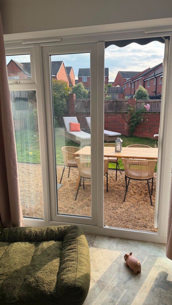 Looking out through glass doors from the Retreat Room onto the secure garden at House of Paws Retreat — a moss-green dog bed indoors, a gravel patio with a wooden dining set, and orange-cushioned chaise lounges on the lawn beyond a brick wall