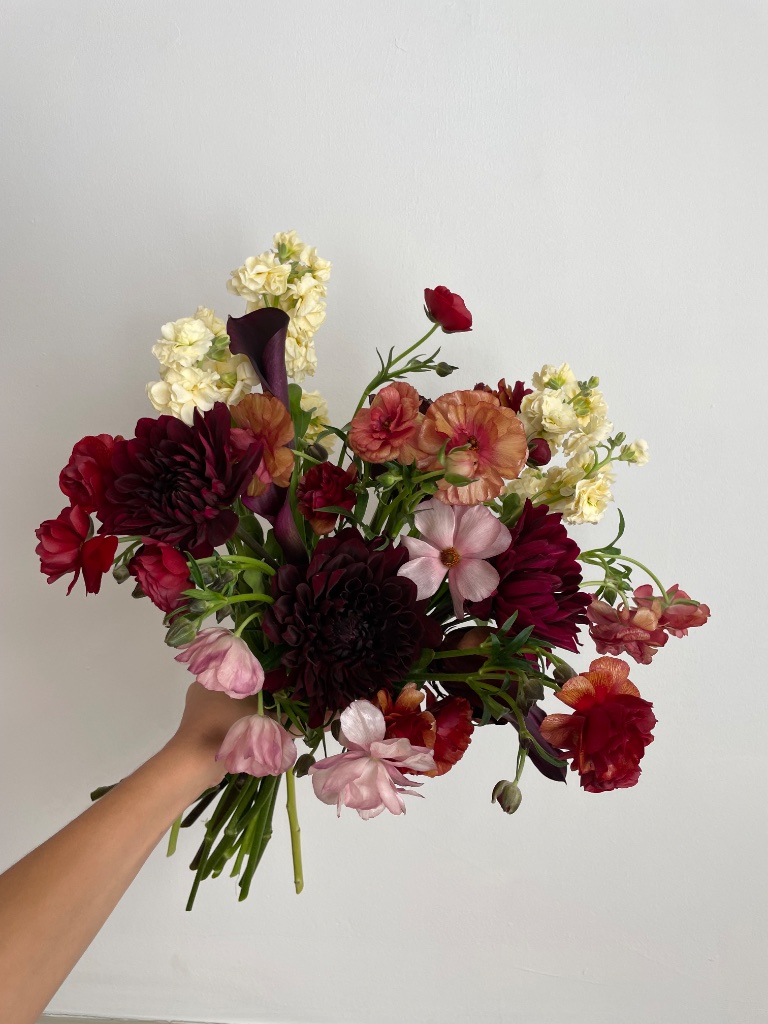 Hand-held bouquet of dahlias, ranunculus and cosmos in deep burgundy tones
