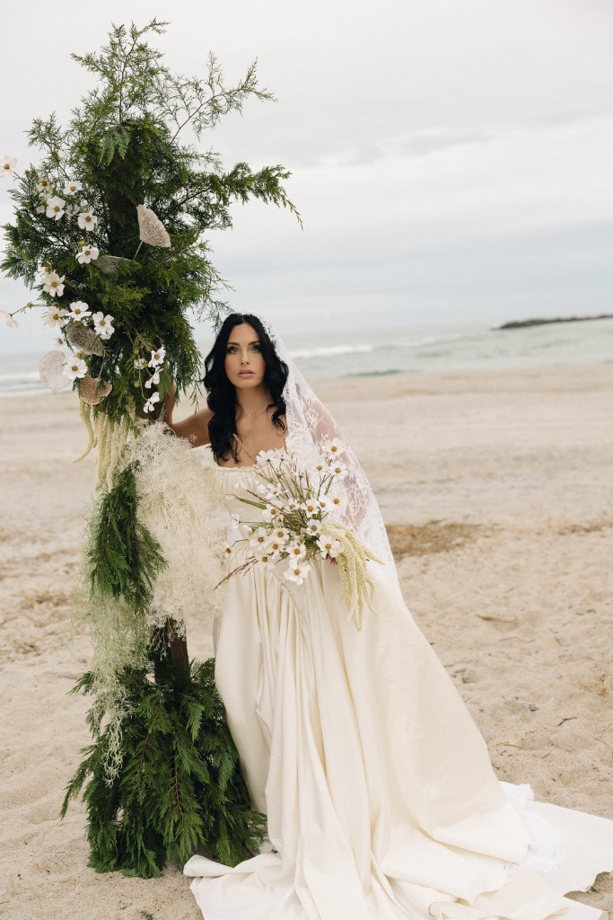 Bride standing beside a tall botanical arch installation on the beach