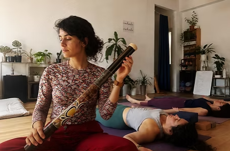 A practitioner holding a didgeridoo during a voice and sound therapy session while participants rest on mats