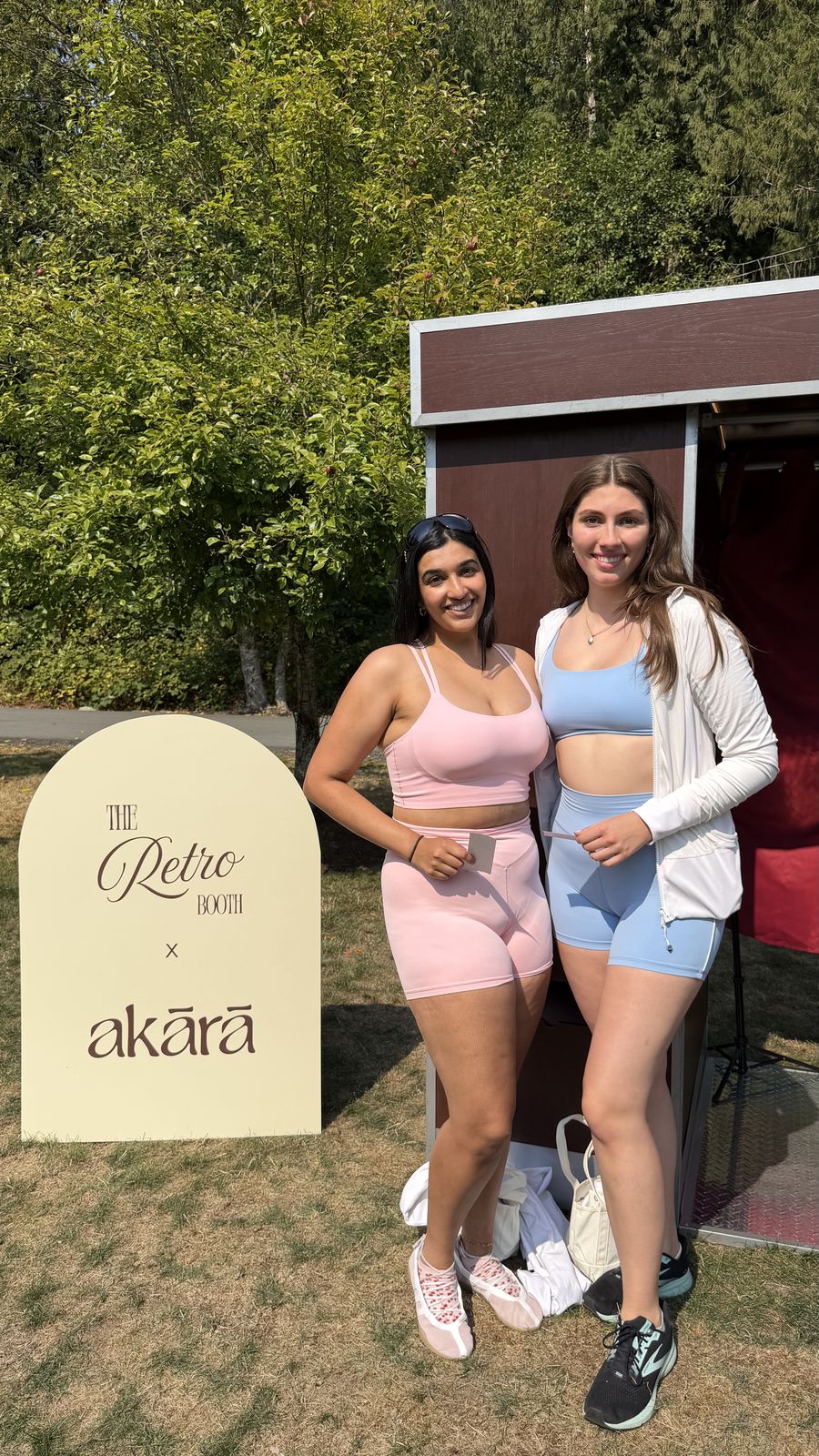 Two guests in activewear posing with photo strips beside the Retro Booth x Akara sign at the Akara Pilates event