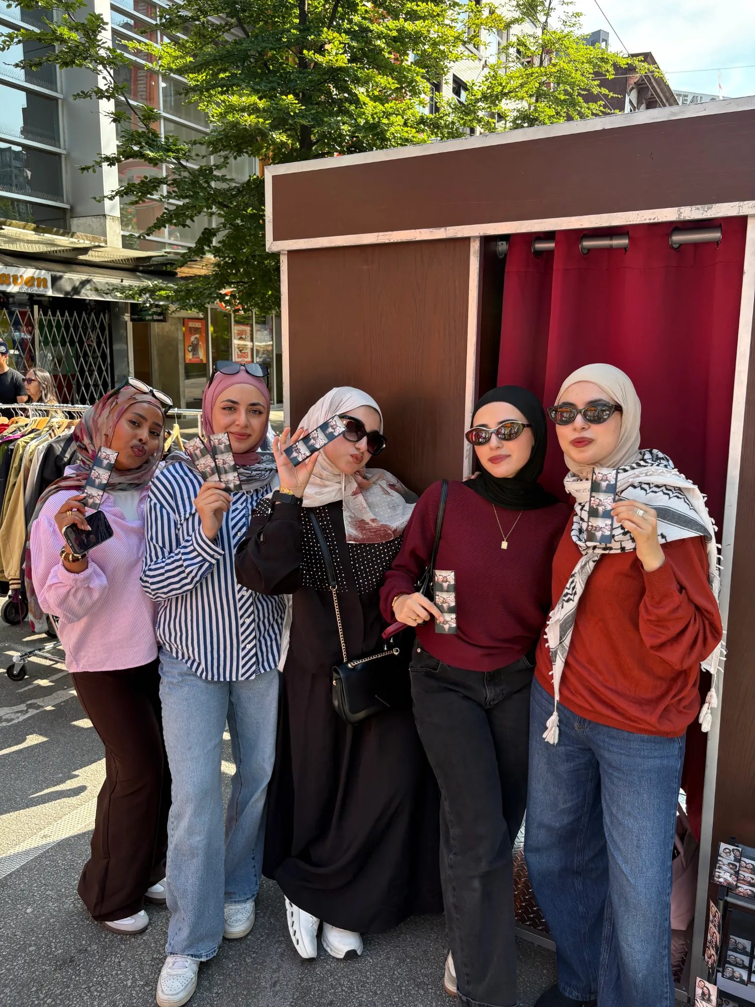 Five friends holding Retro Booth photo strips in front of the Snap a Core Memory backdrop at a Vancouver street festival