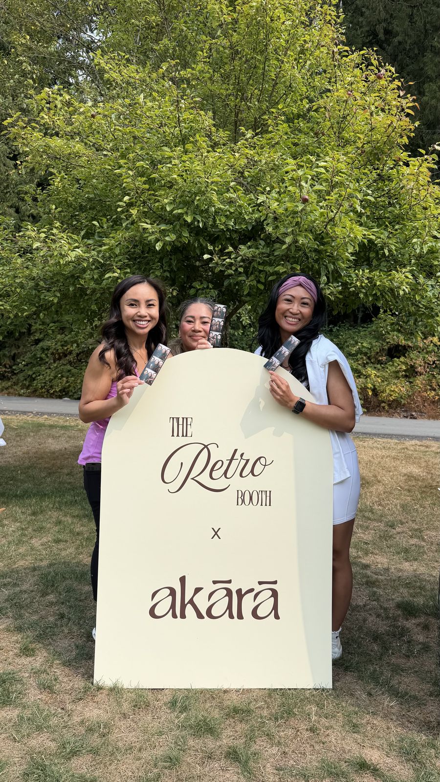 Three guests smiling and holding photo strips behind the Retro Booth x Akara sign at the Akara Pilates event