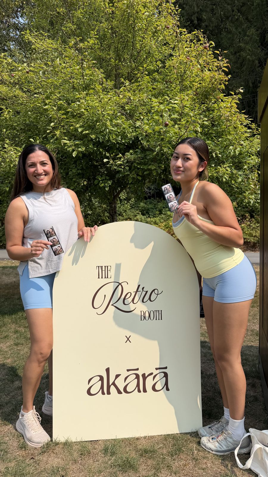 Two guests posing with photo strips beside the Retro Booth x Akara sign at the Akara Pilates event in Langley