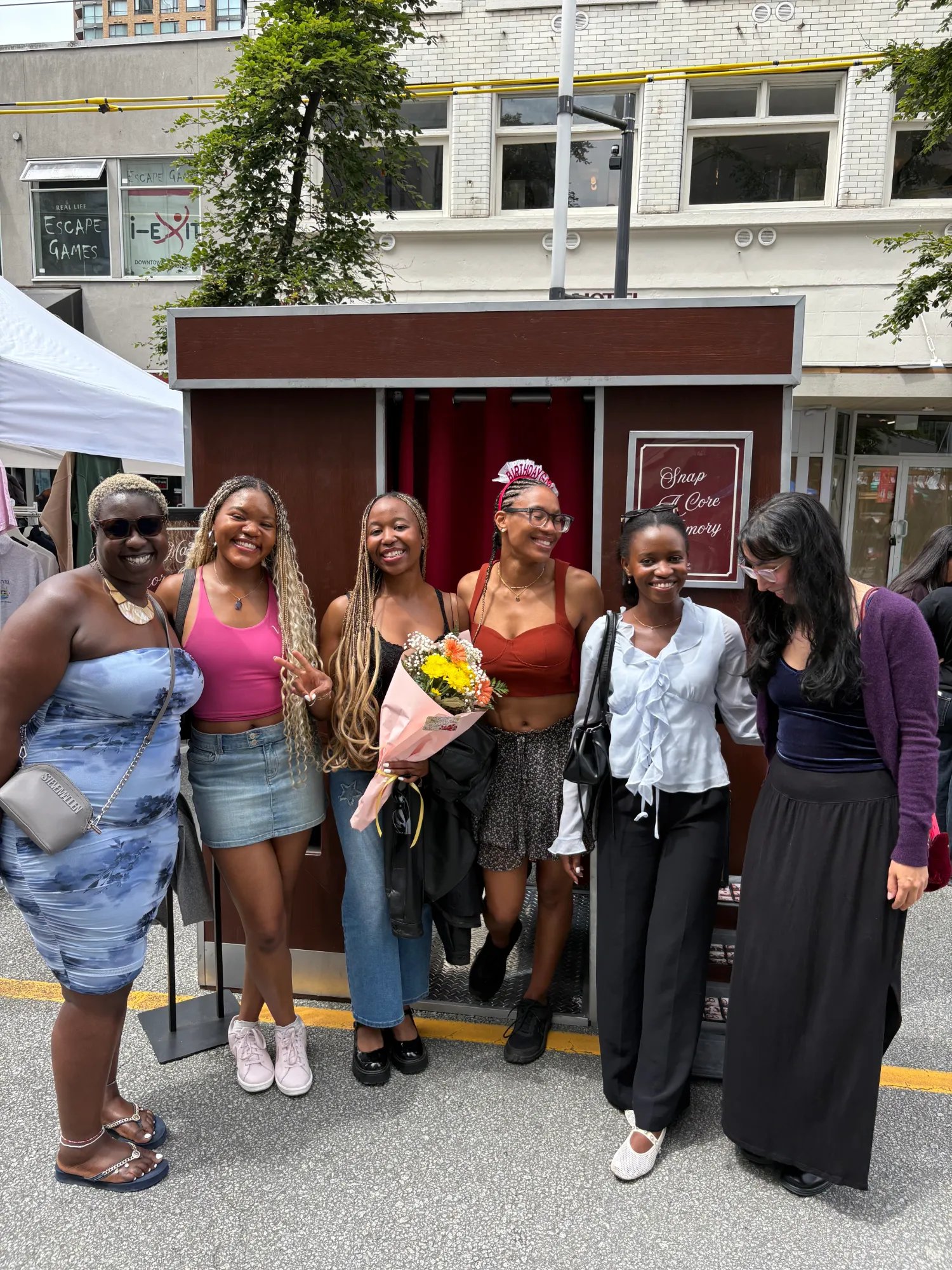A group of friends holding Retro Booth photo strips in front of the Snap a Core Memory backdrop in Vancouver