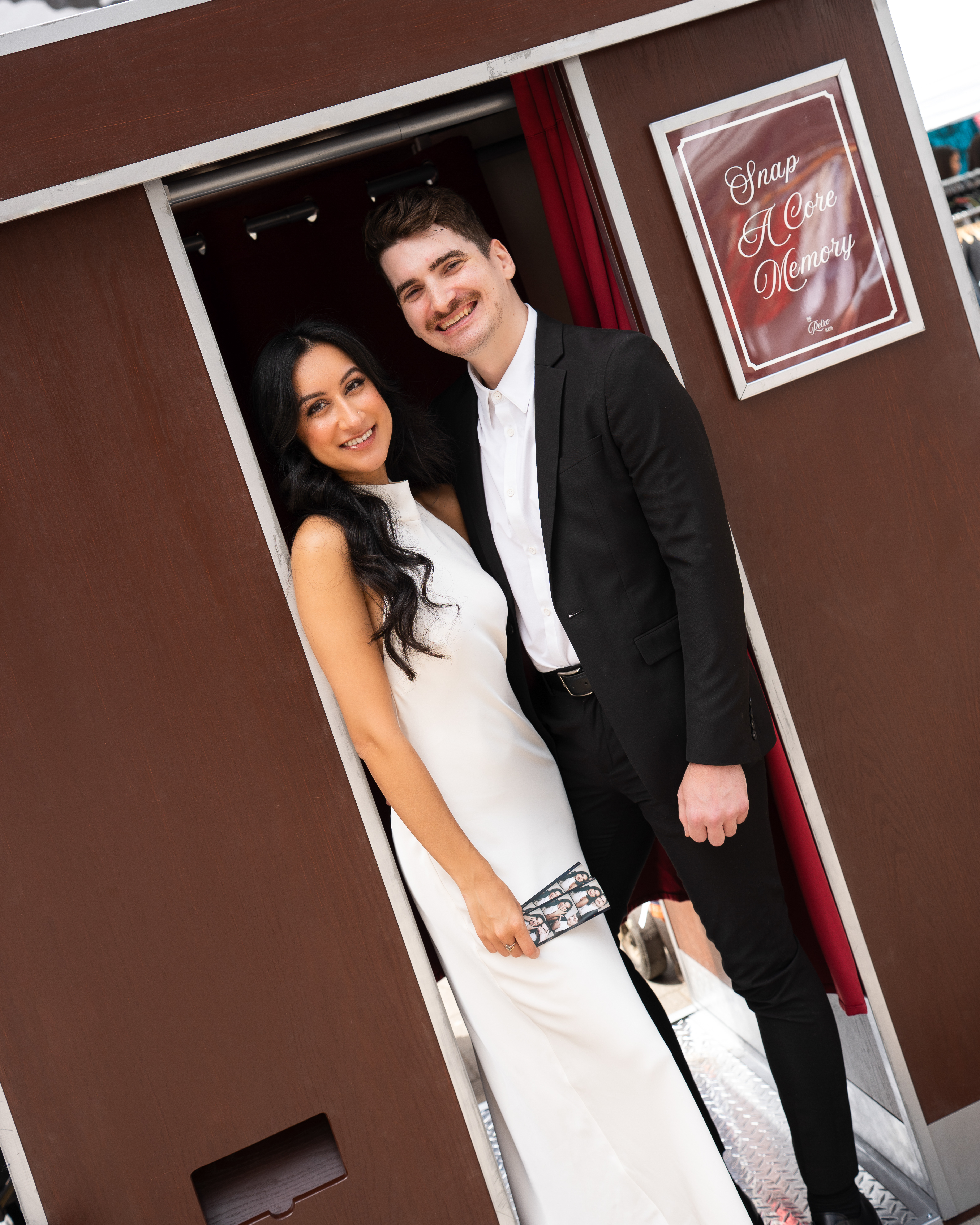 A couple smiling in the doorway of The Retro Booth's enclosed vintage photo booth, holding a printed photo strip