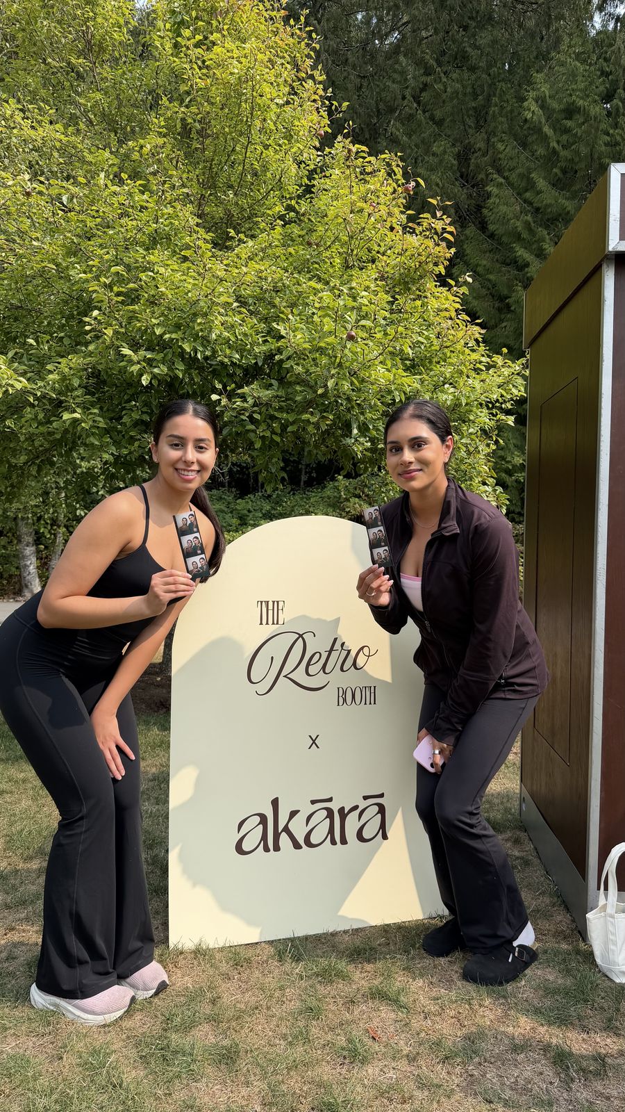 Two guests in workout attire holding Retro Booth photo strips beside the Retro Booth x Akara sign at the Akara Pilates event in Langley