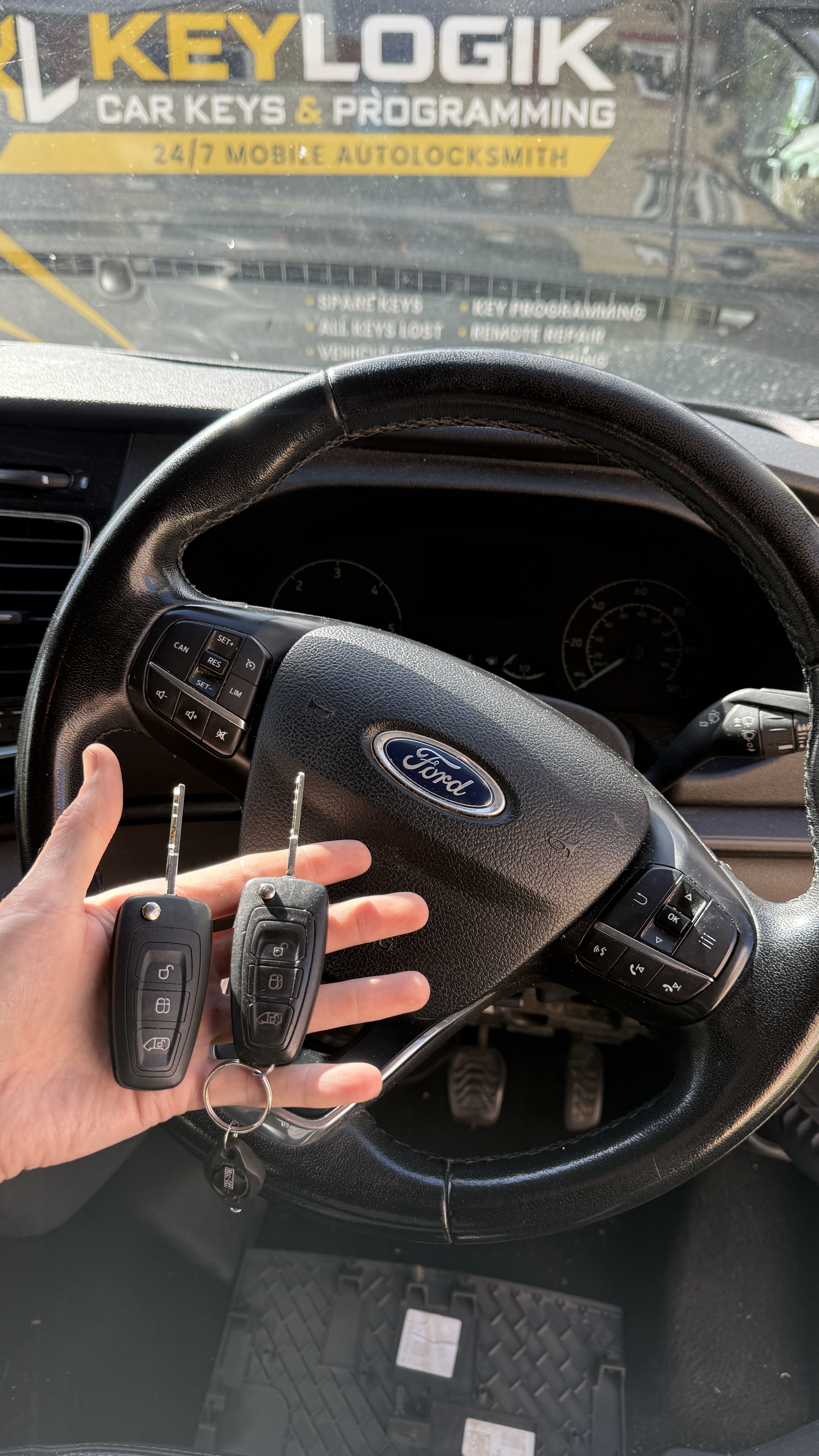Two Ford keys held at the steering wheel with KeyLogik mobile auto locksmith signage visible through the windscreen