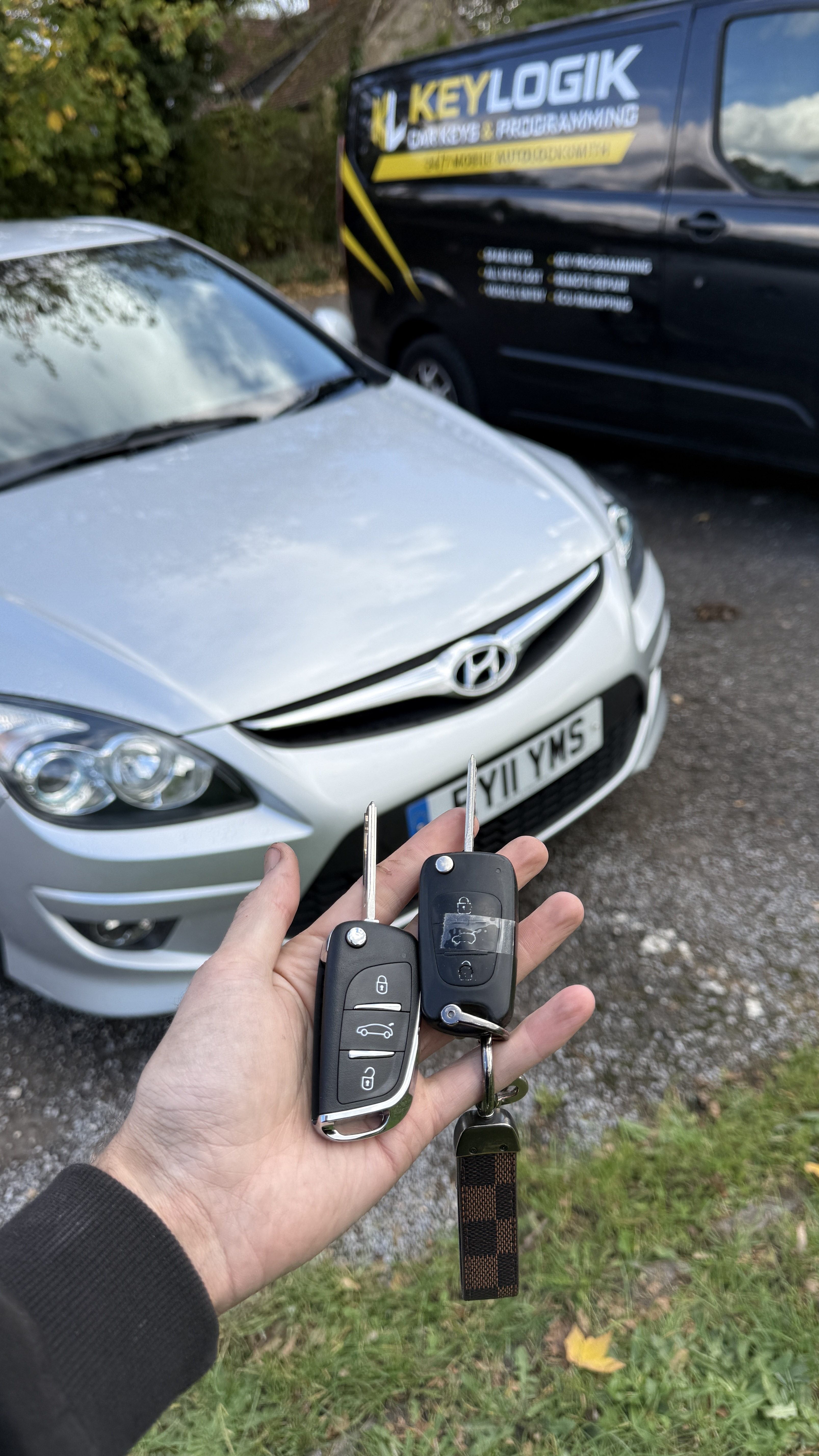 New and original flip keys held in front of a silver 2011 Hyundai i30 beside the KeyLogik van after spare key programming in Guildford, Surrey