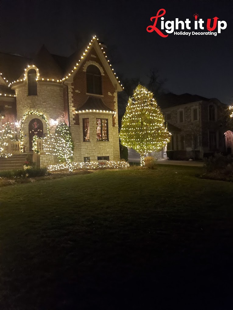 Elegant stone home with warm white roofline lights and lit evergreen trees