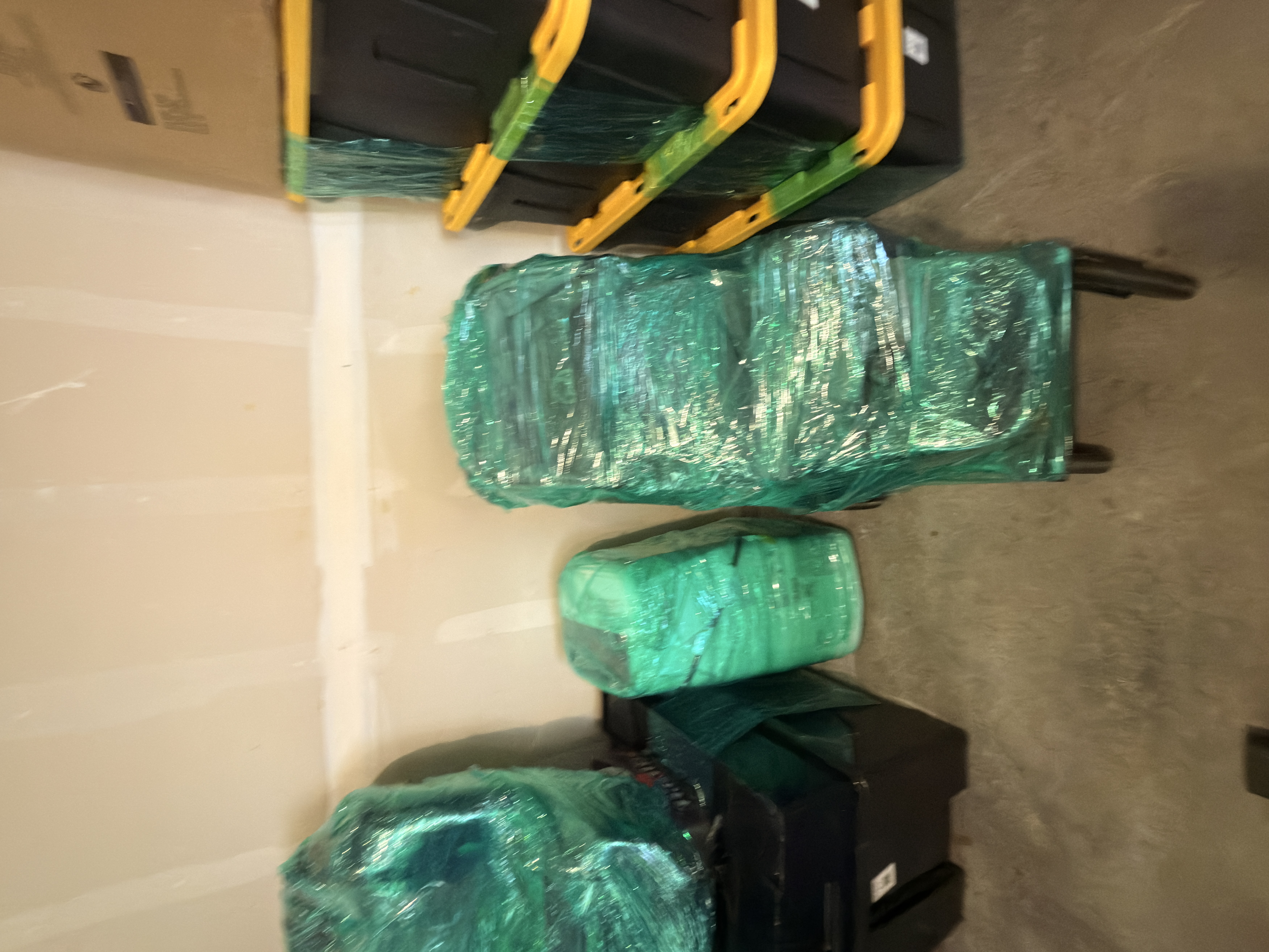 Several items tightly wrapped in translucent green plastic stretch wrap, with black and yellow storage totes behind them.