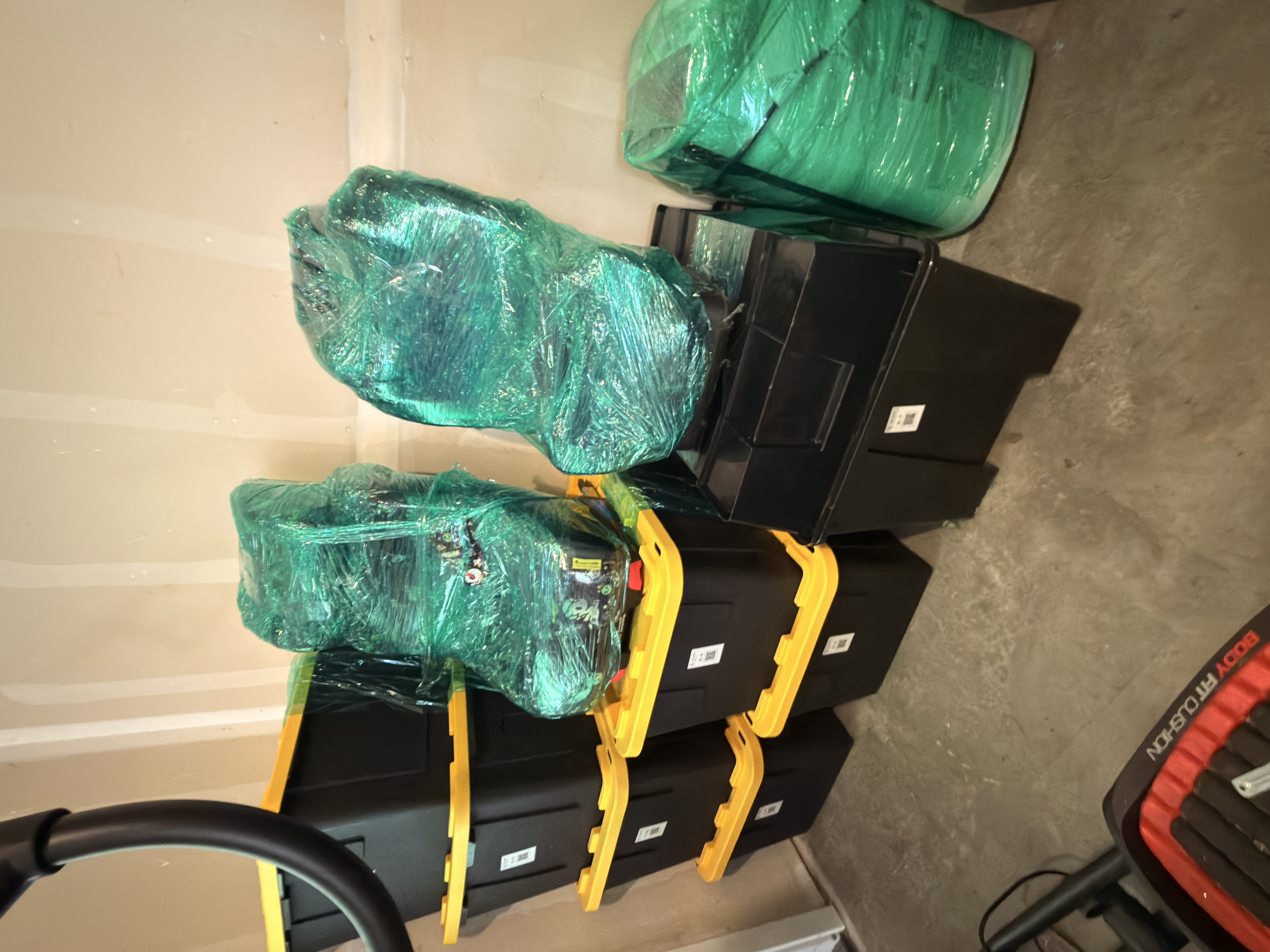 Black storage totes with yellow lids and QR code labels, with bulky items wrapped in green plastic film on top.