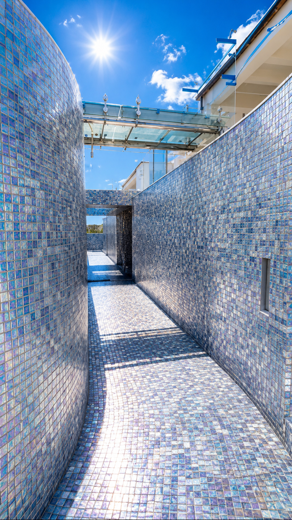Iridescent blue glass mosaic corridor with mosaic-clad walls and floor beneath a glass canopy, opening to the sunlit sea beyond