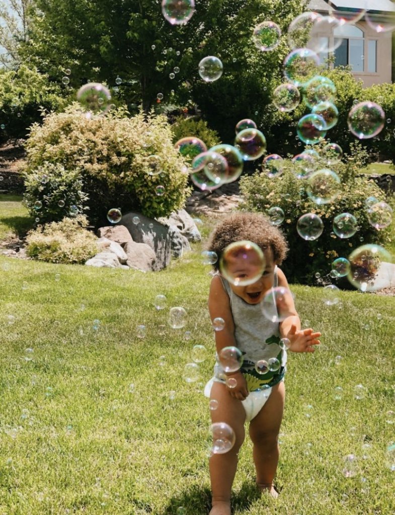 A toddler laughing and reaching for bubbles in a grassy backyard