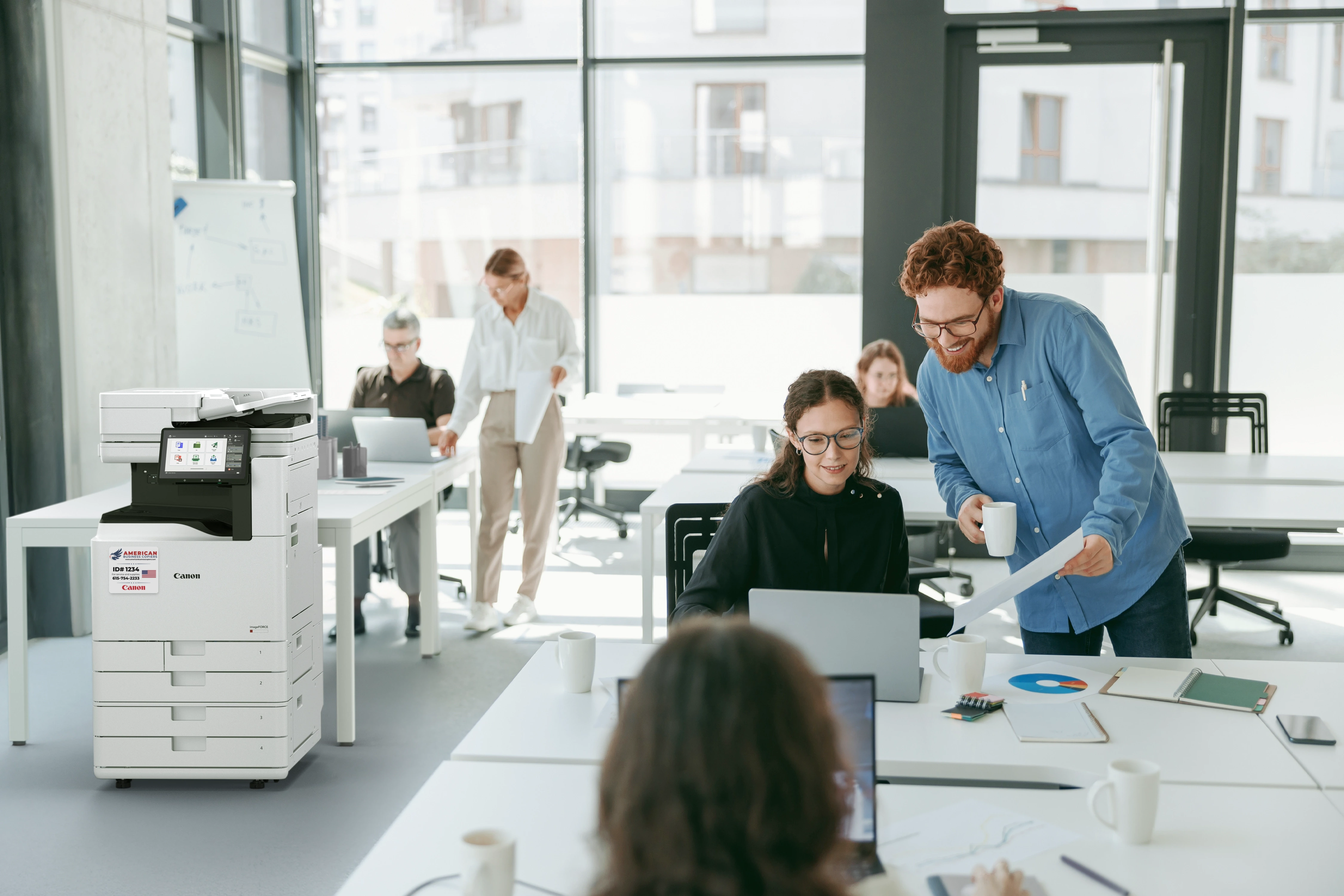 A modern office team collaborating around a Canon imageFORCE multifunction printer in a bright, daylit workspace
