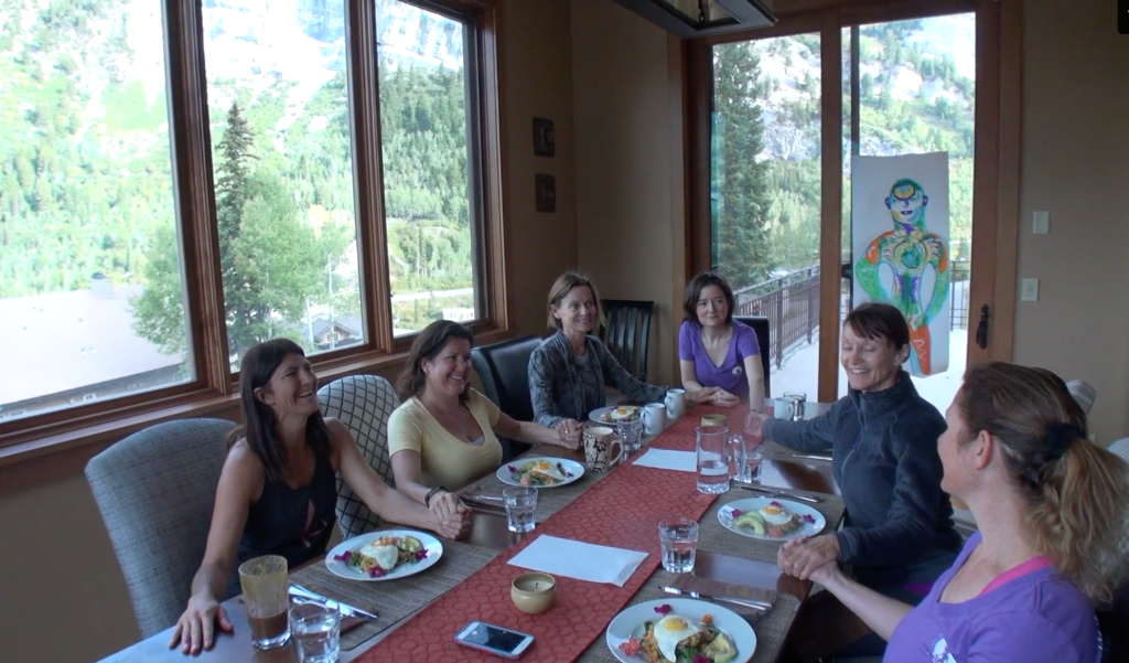 Women laughing and sharing a meal together at a dining table with mountain views