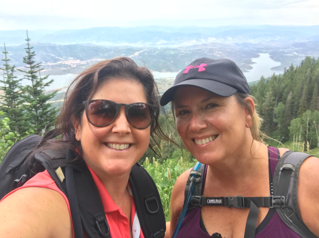 Two women smiling at summit with lake and mountain valley view behind them