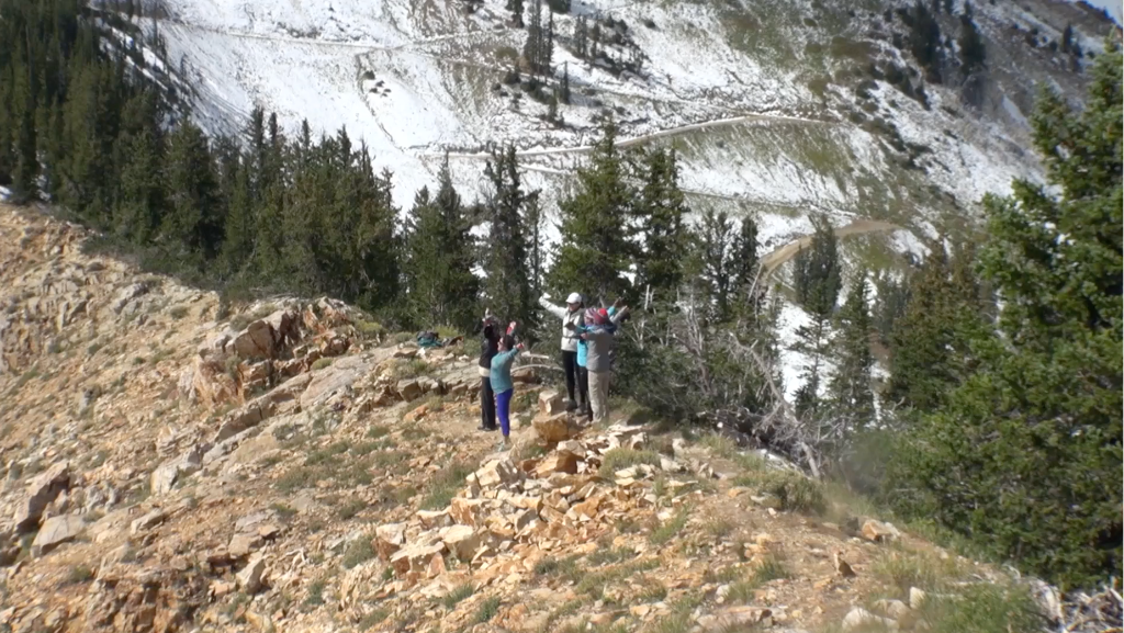 Group of women with arms raised on a rocky mountain ridge with snow-capped peaks behind