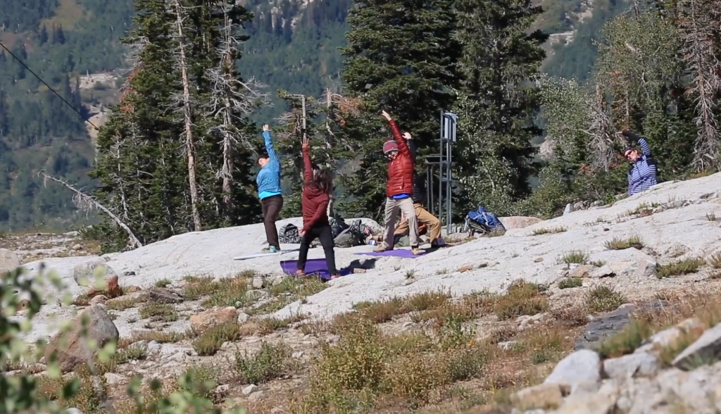 Group doing yoga poses on a rocky alpine slope surrounded by pine trees