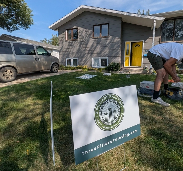 A painter from 3 Pillars Painting working on the exterior of a grey residential house with a yellow front door, featuring a company yard sign in the foreground