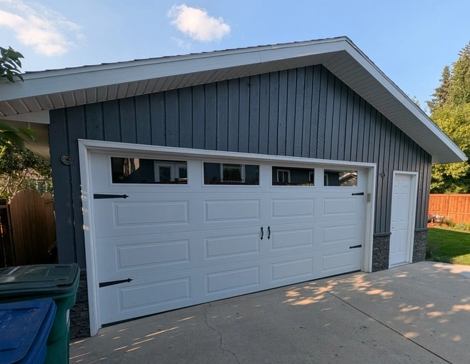 Renovated residential detached garage featuring dark grey vertical siding, white carriage-style door with windows, and grey stone veneer base