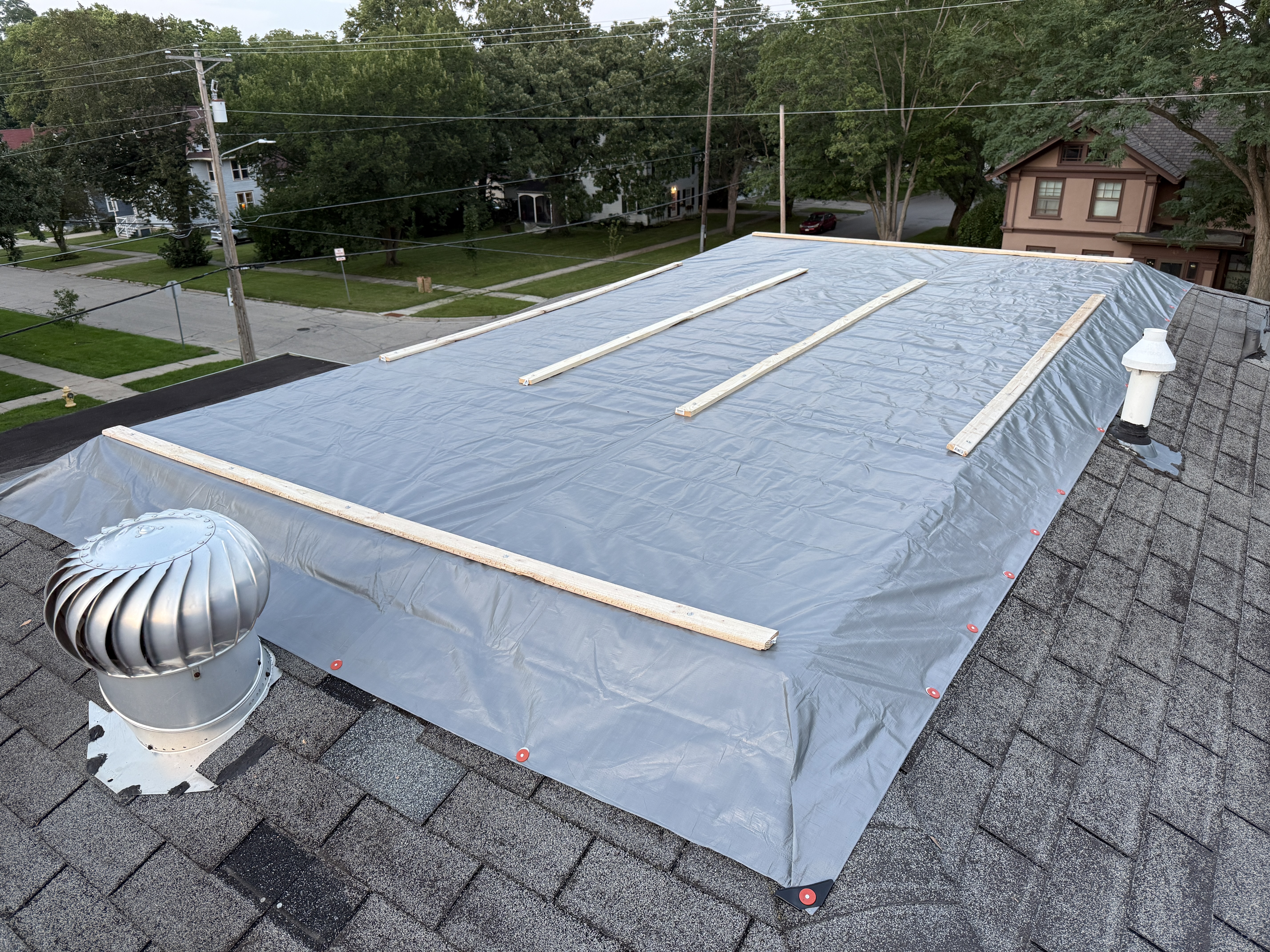 Grey tarp secured over the roof repair area with wooden furring strips and red fastener caps