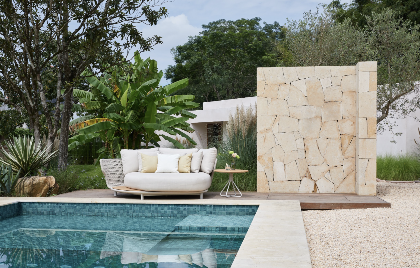 Daybed Alma en acabado Beige sobre terraza de madera junto a alberca con muro de piedra