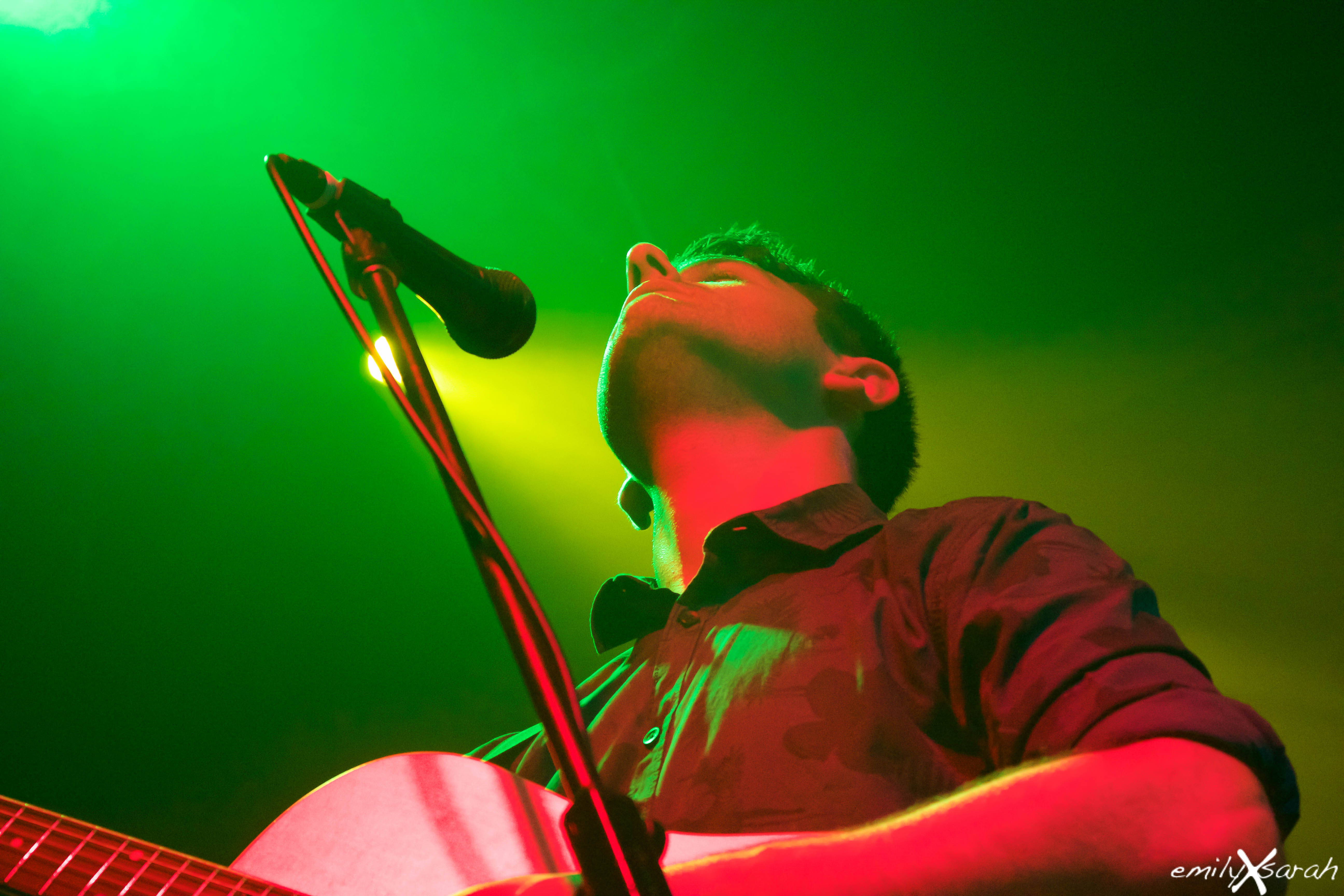 Michael Pace singing into a microphone while playing acoustic guitar on stage
