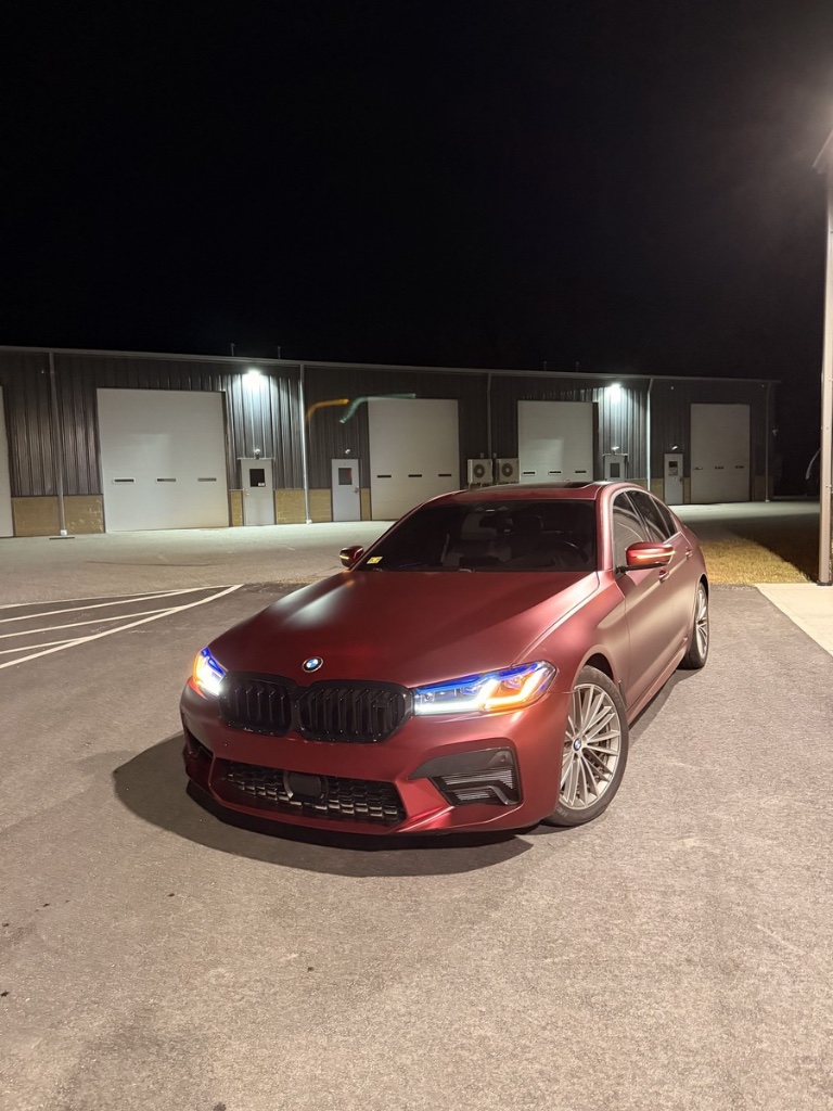 BMW 5 Series sedan wrapped in a satin red color change wrap by Xtreme Graphics Design in Rhode Island, photographed at night in front of a warehouse, after the vehicle wrap installation.
