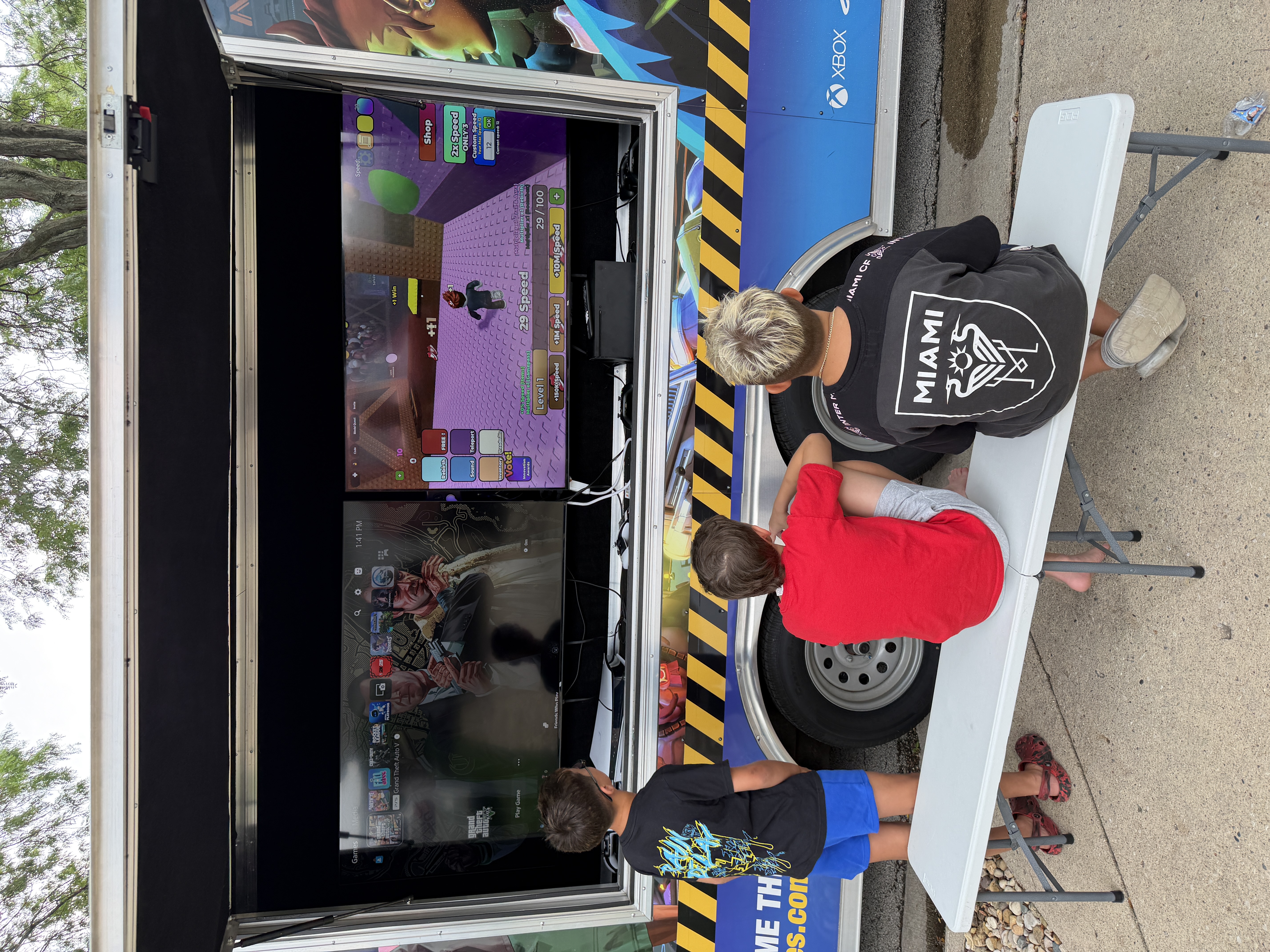 Three boys playing video games on dual stacked monitors inside the GameRush gaming truck, with an Xbox dashboard and GTA V visible on screen