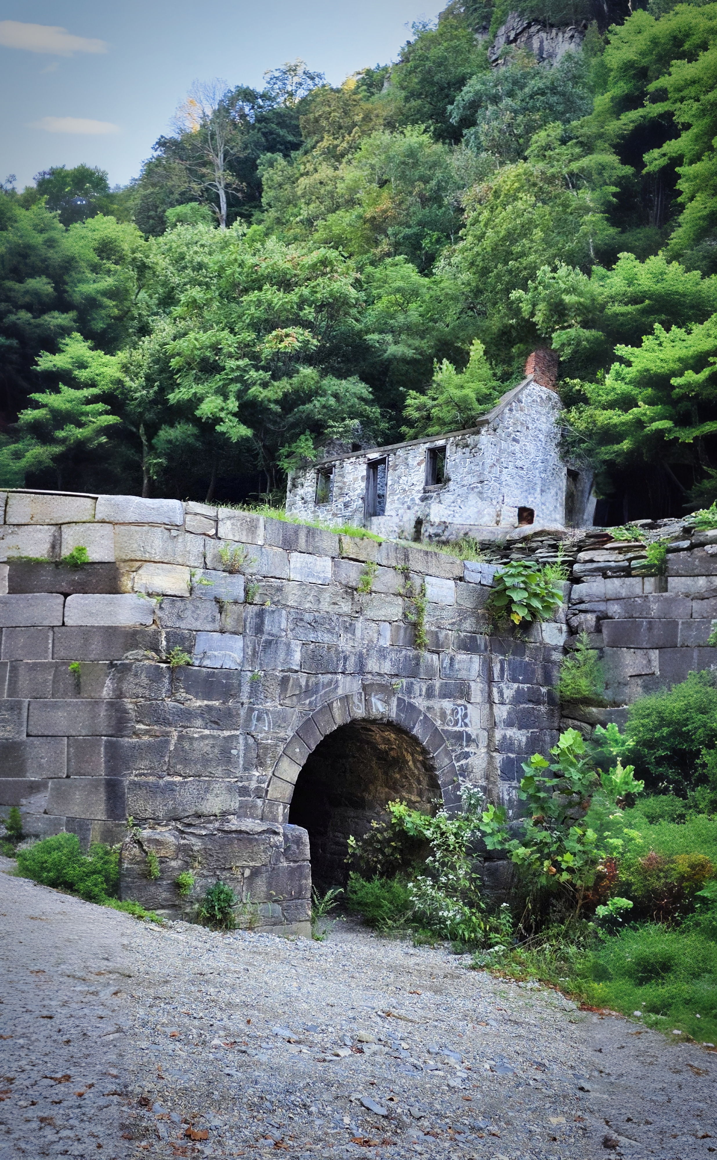 A stone culvert along the towpath near Harpers Ferry — the canal's masonry still holding its arch, with the roofless ruin of a stone house and its brick chimney reclaiming to the woods behind it.