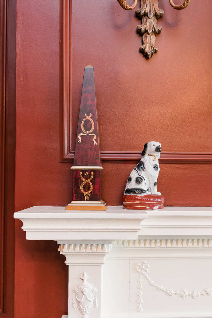 Detail of the study mantel — a red obelisk and a porcelain spaniel flank the carved frieze, with a wall sconce above.
