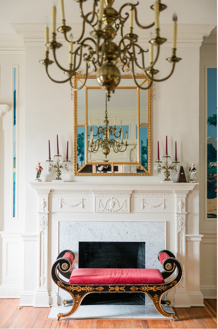 The mantel in the Lafayette Parlor — carved swags, dentil molding, and a gold-framed mirror above, flanked by landscape murals of rural life.