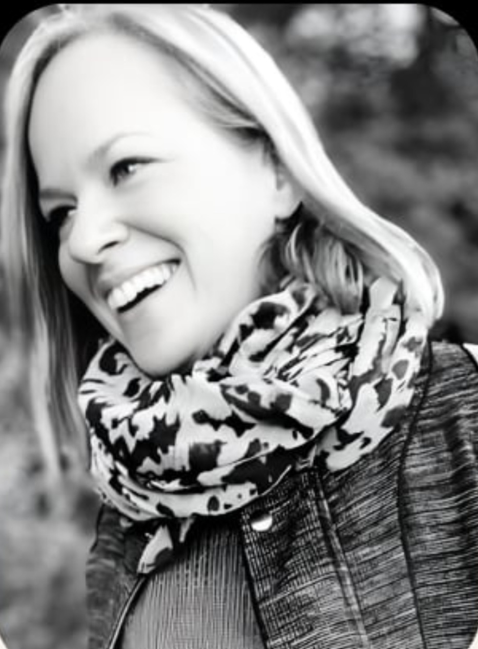 A black-and-white photo of Ros, smiling and looking off to the left, blonde hair tucked behind her ears, wearing a patterned scarf and a jacket, against a soft out-of-focus background