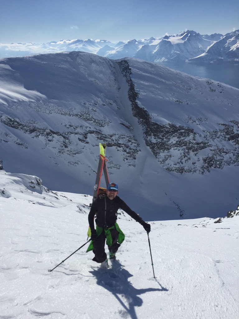 Backcountry skier climbing steep Lyngen Alps face with fjord and peaks behind