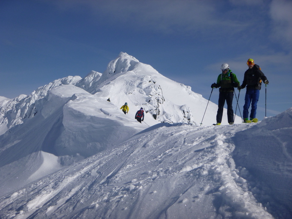 Four backcountry skiers traversing high-altitude snow ridge in Lyngen Alps, Norway, with prominent snow-capped peak behind