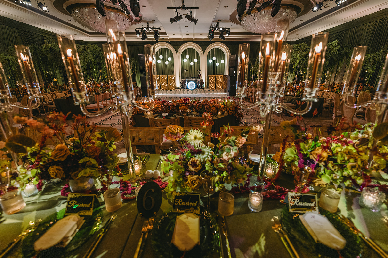 Elegant wedding reception banquet hall with formal table settings, crystal candelabras, floral centerpieces, and a live band stage with warm ambient lighting. Live Pros wedding AV and production in California.