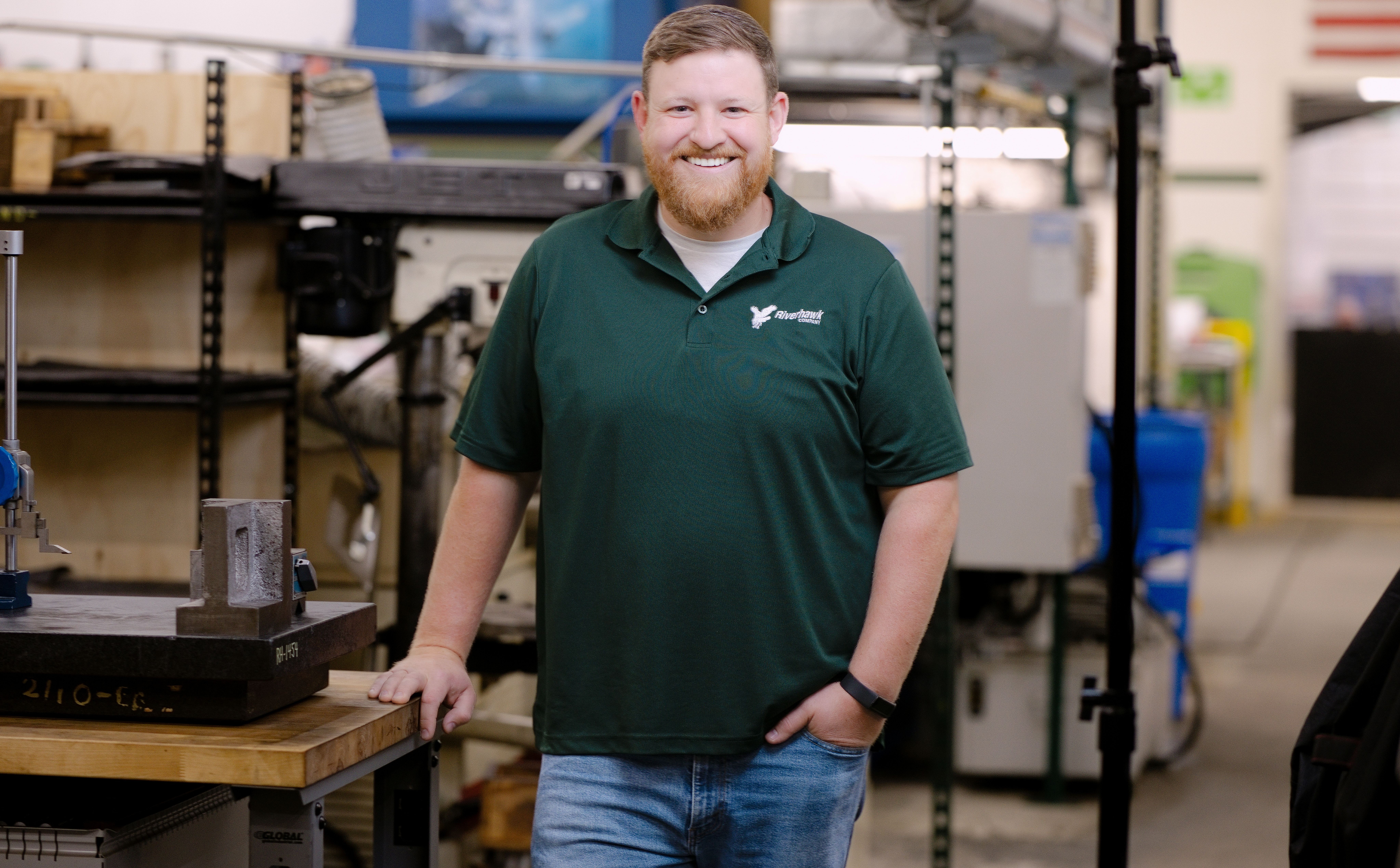 Spencer Roose wearing a Riverhawk Company polo shirt, standing in an industrial machine shop