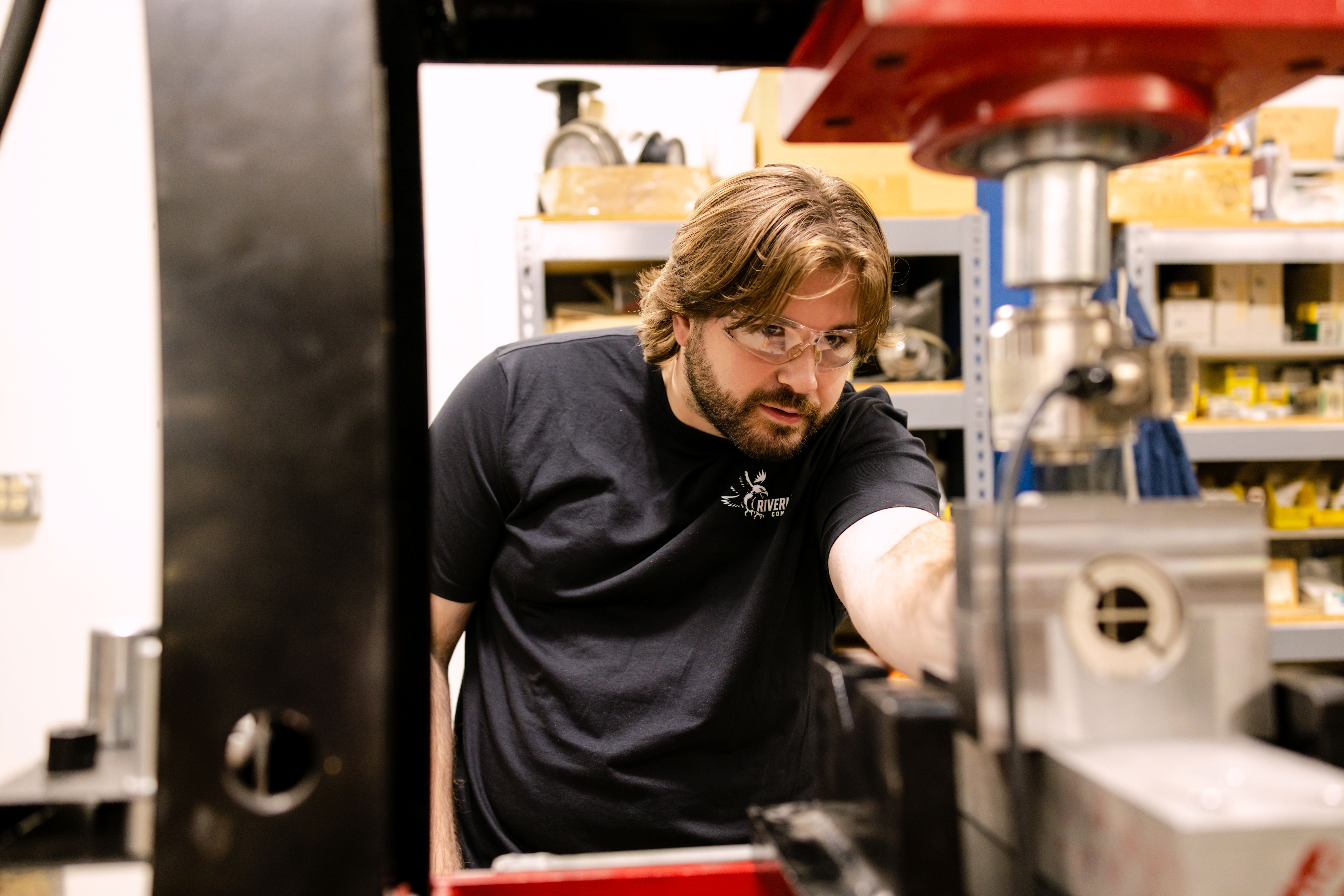 Dawson Kiser wearing safety glasses and a Riverhawk logo t-shirt, inspecting industrial machinery in a workshop setting