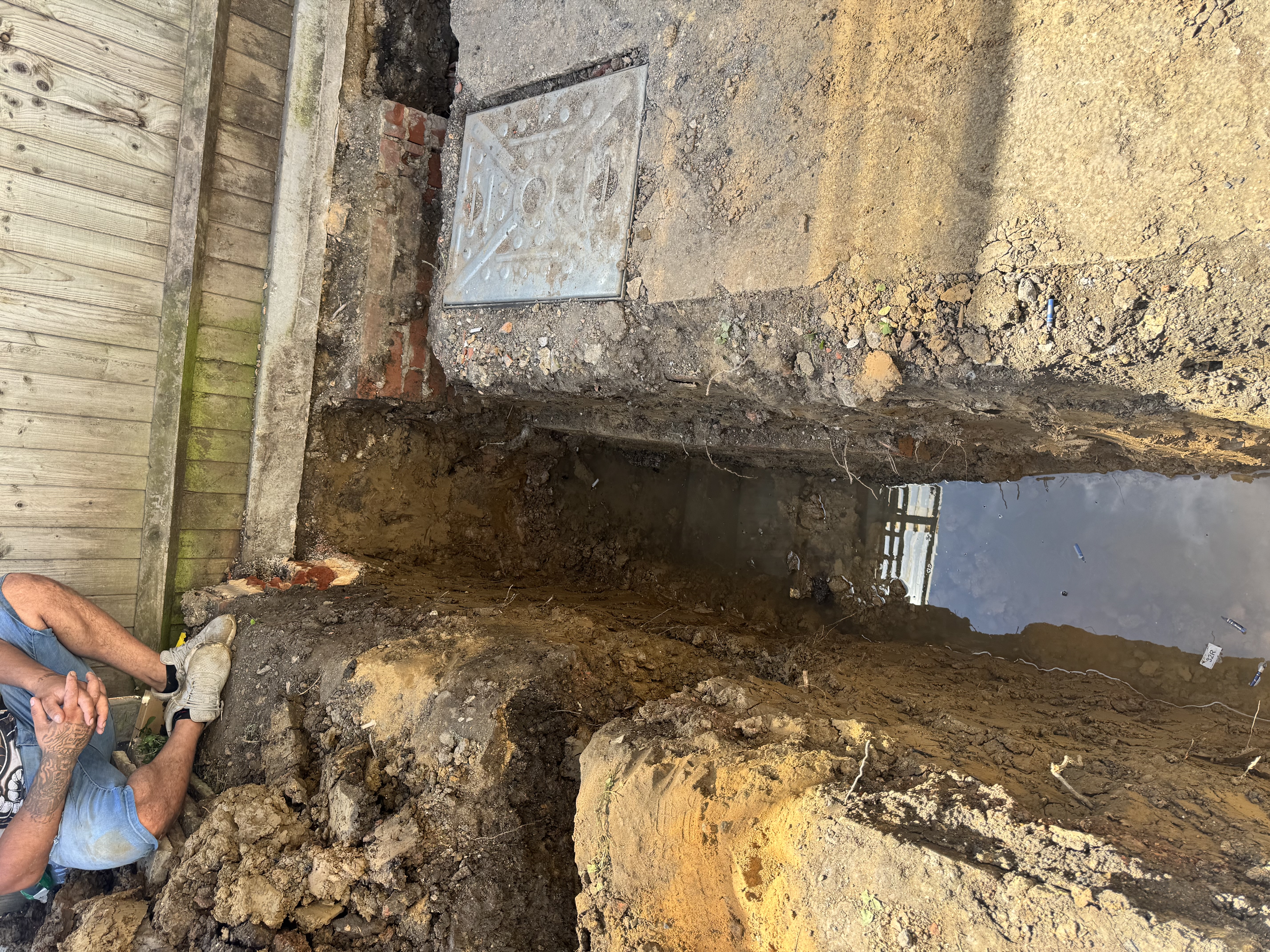 Open trench in a garden showing soil layers and a drainage access cover