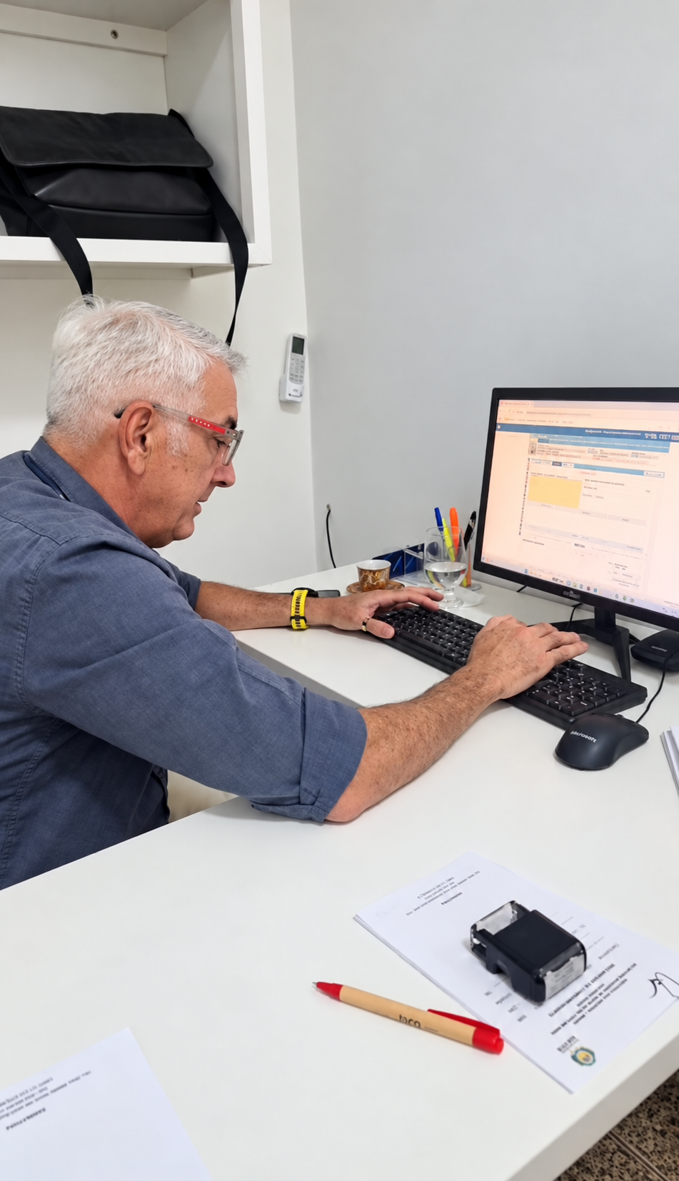 Profissional trabalhando no computador em ambiente de consultório, com documentos e carimbo sobre a mesa