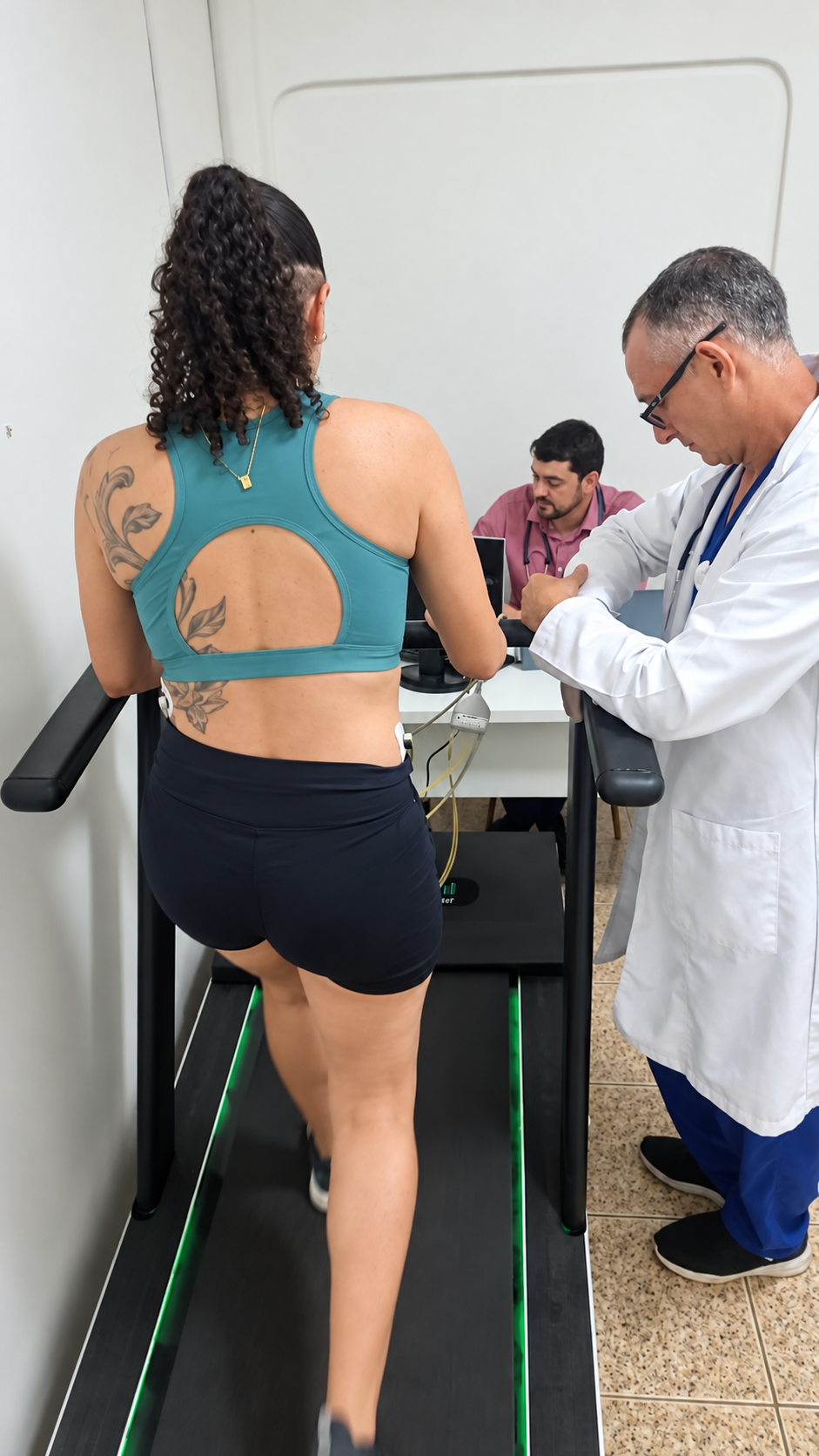 Paciente caminhando em esteira com sensores de ECG no dorso, acompanhada por profissional de saúde de jaleco branco