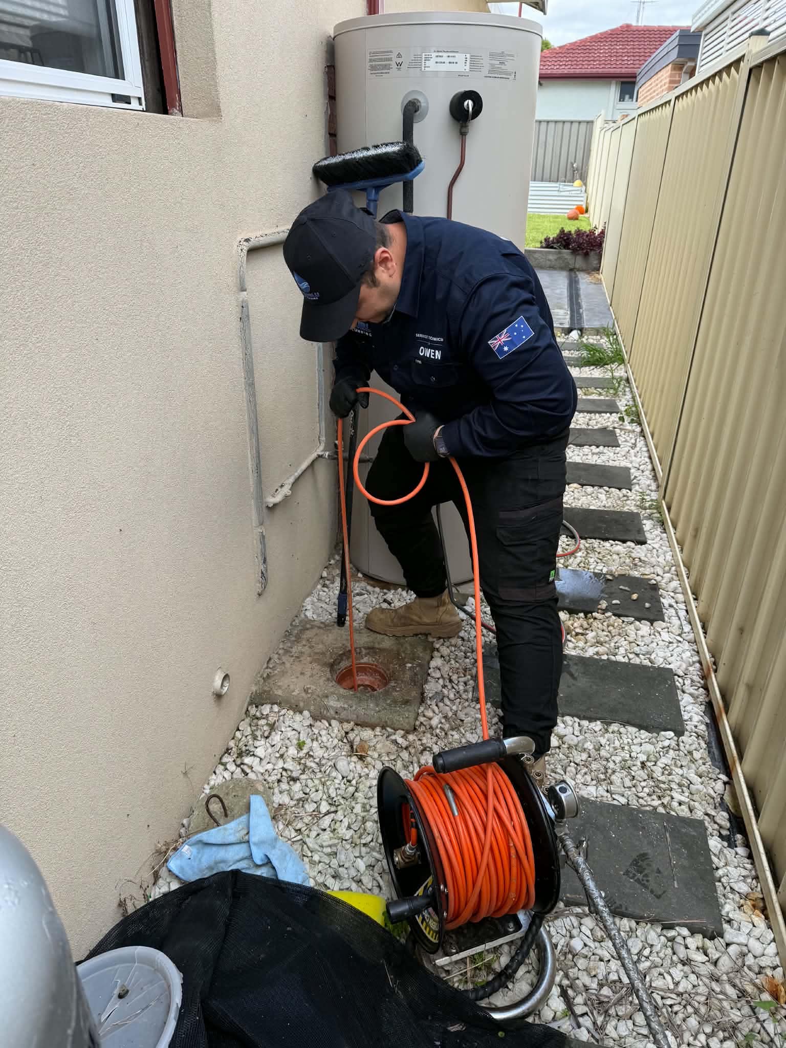 Tru Blu service technician feeding a drain inspection cable into an outdoor access point beside a water heater