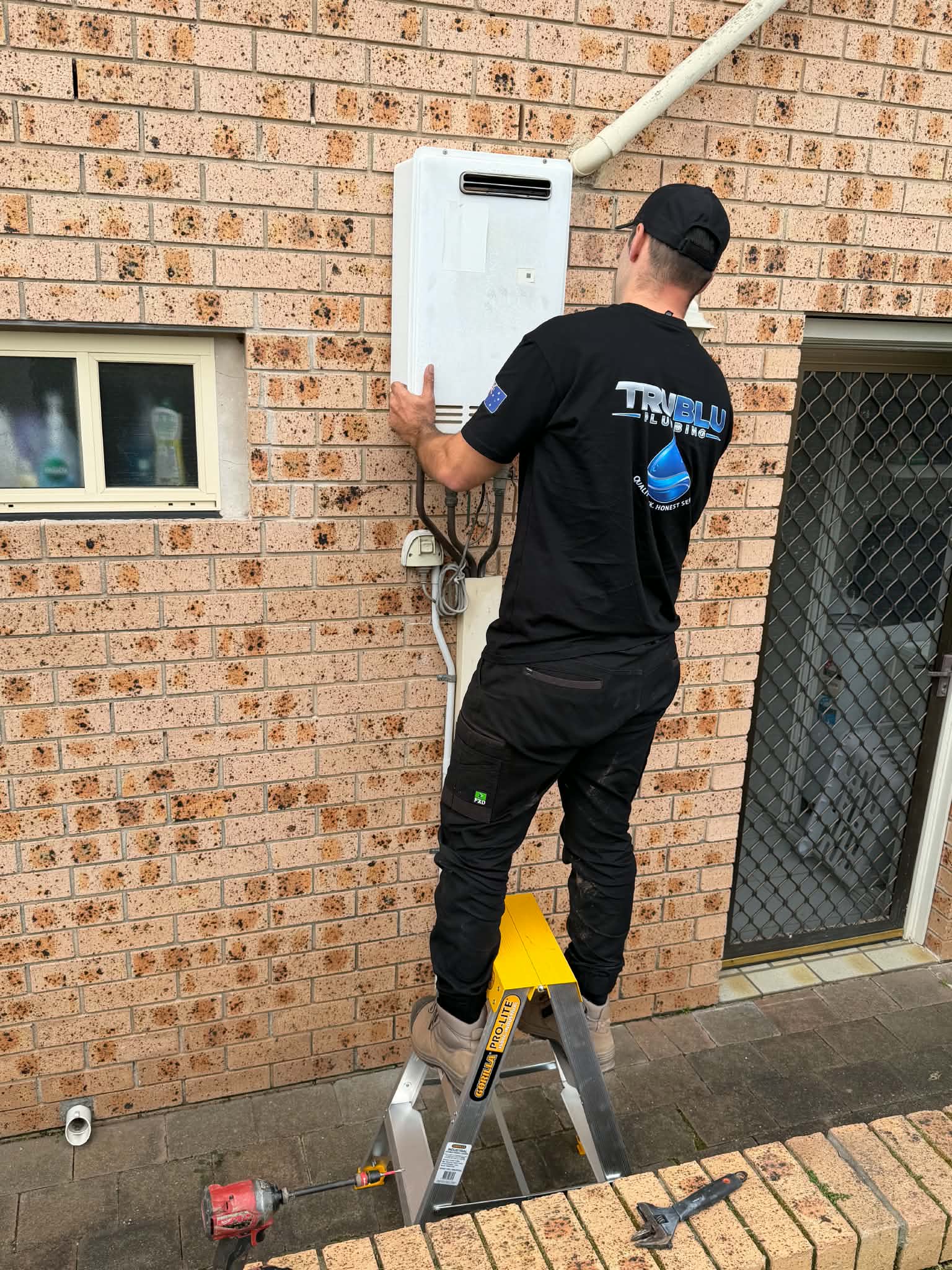 Tru Blu plumber on a ladder installing a wall-mounted gas heater unit against a brick wall