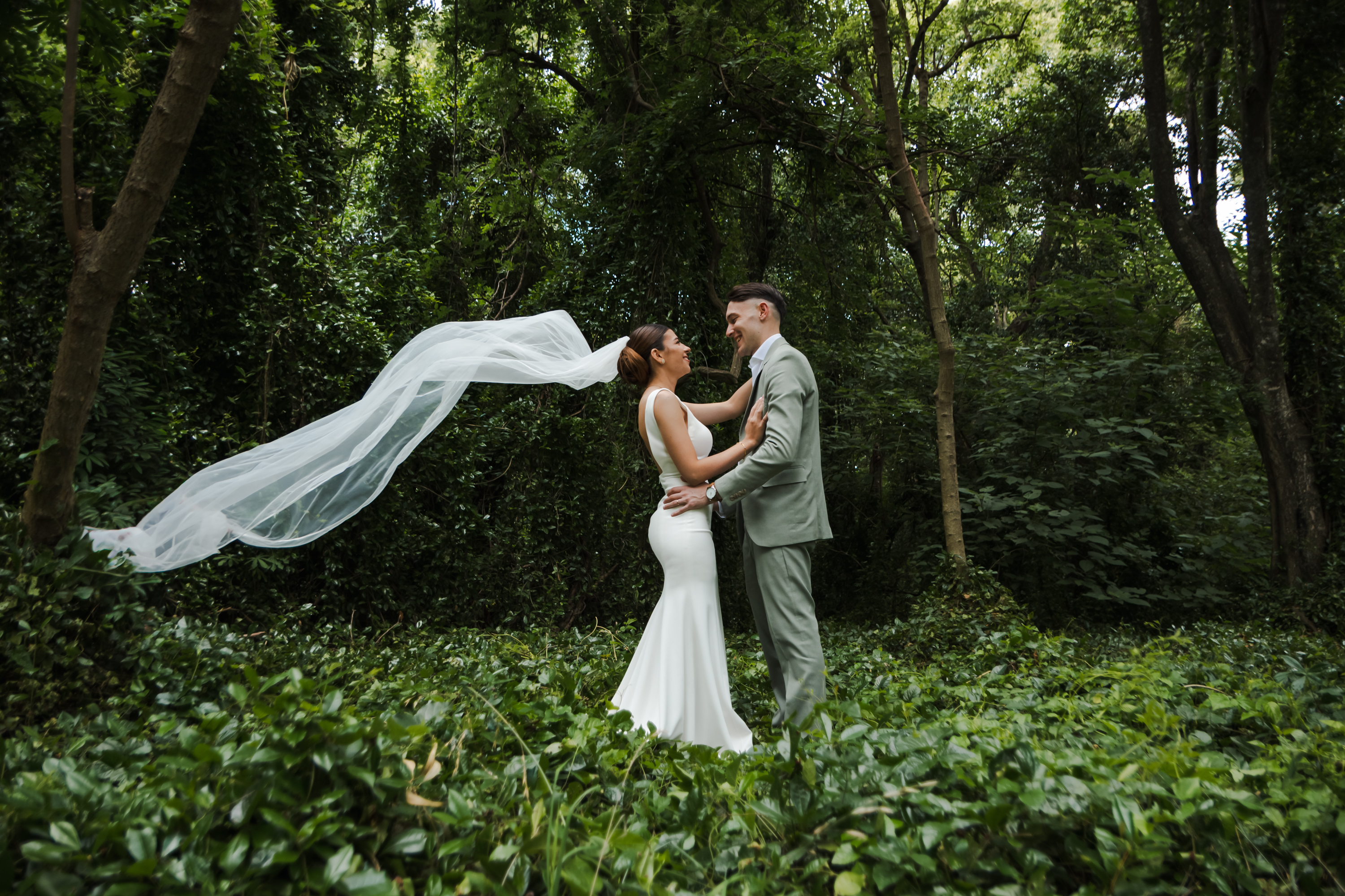 Novios frente a frente en el bosque con el velo de la novia flotando en el aire