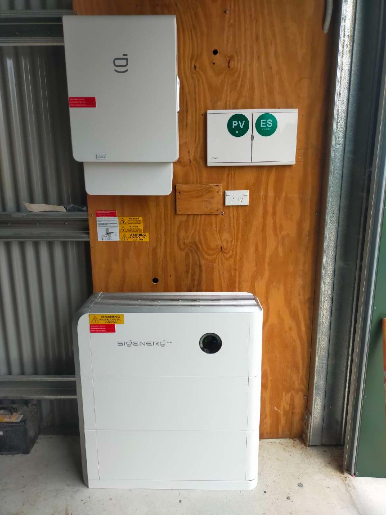 Wall-mounted Sigenergy hybrid inverter, backup gateway enclosure and floor-standing battery unit installed on a plywood-backed wall