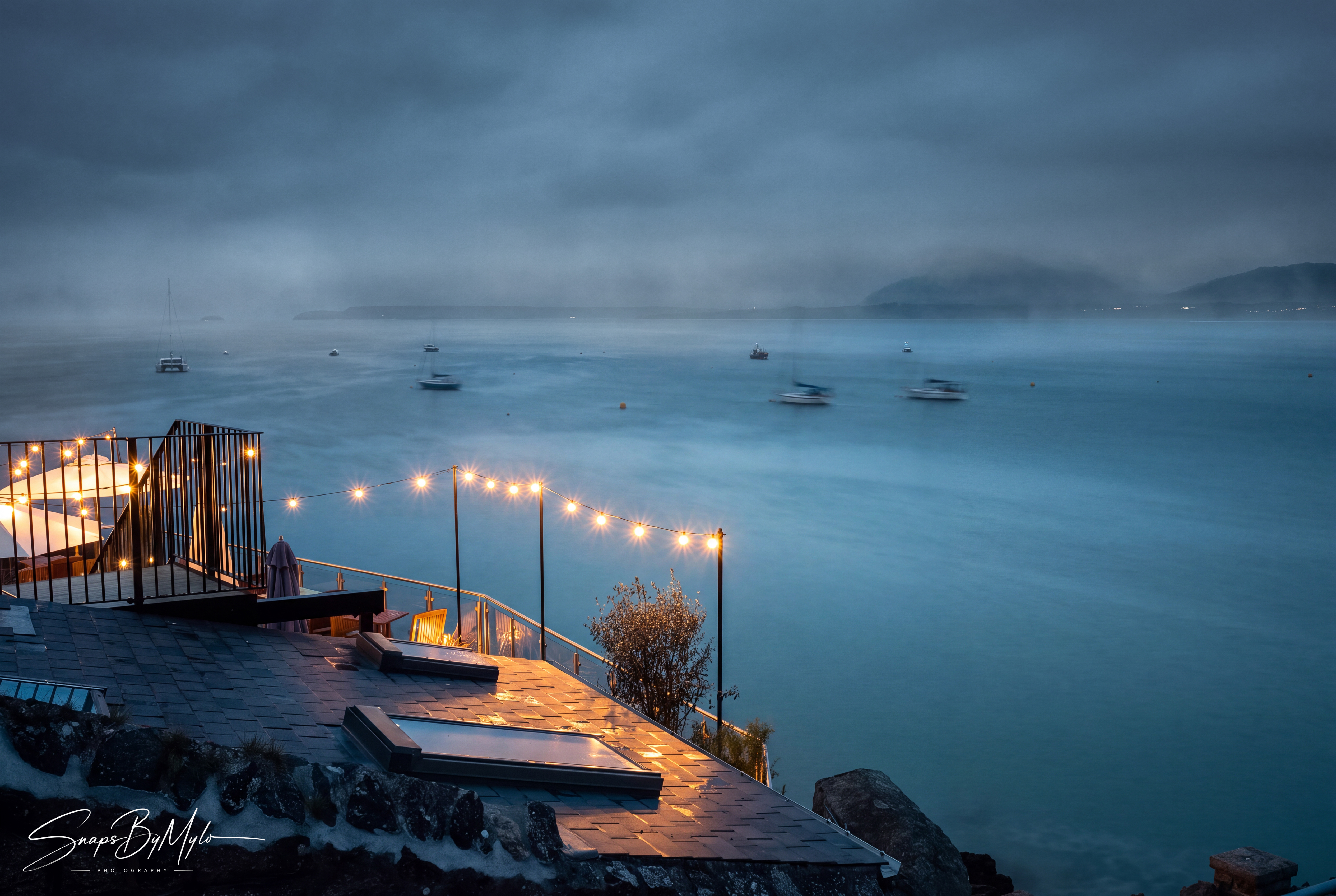 A coastal terrace at twilight, string lights glowing against a moody blue hour sky and sea