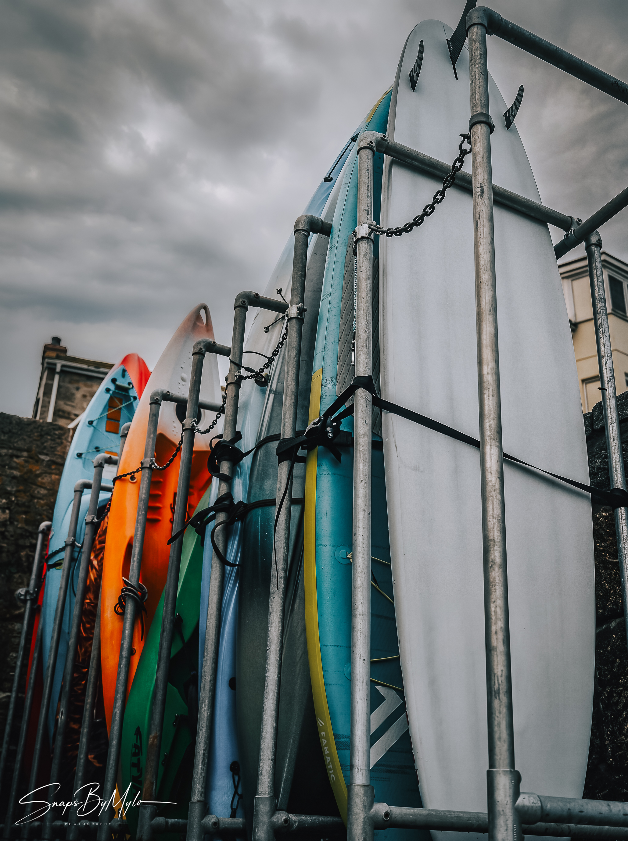 Surfboards and kayaks stored in an outdoor metal rack under a heavy, overcast sky