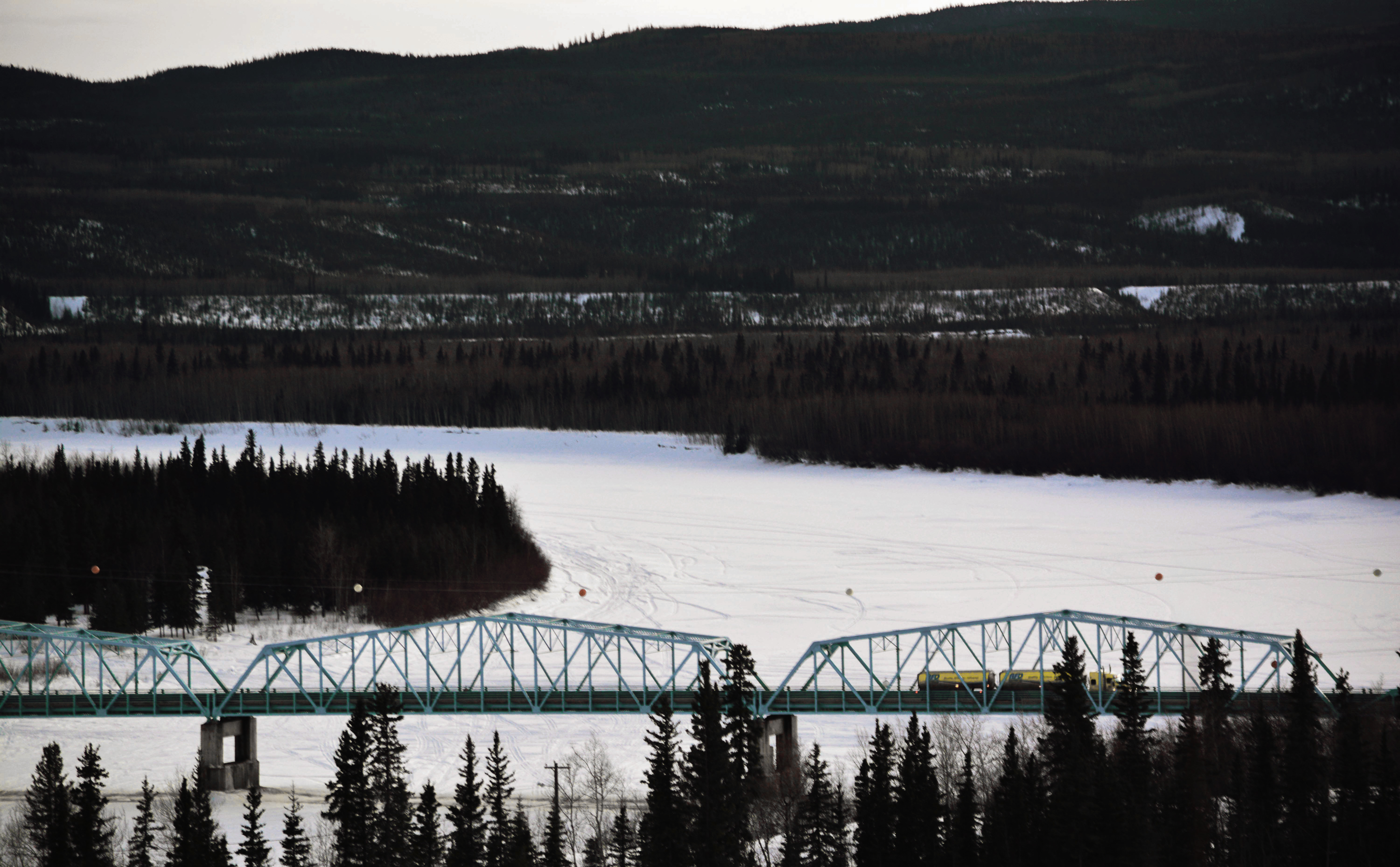 High-angle dusk view of a green truss bridge crossing a frozen river with a yellow AFD tanker truck