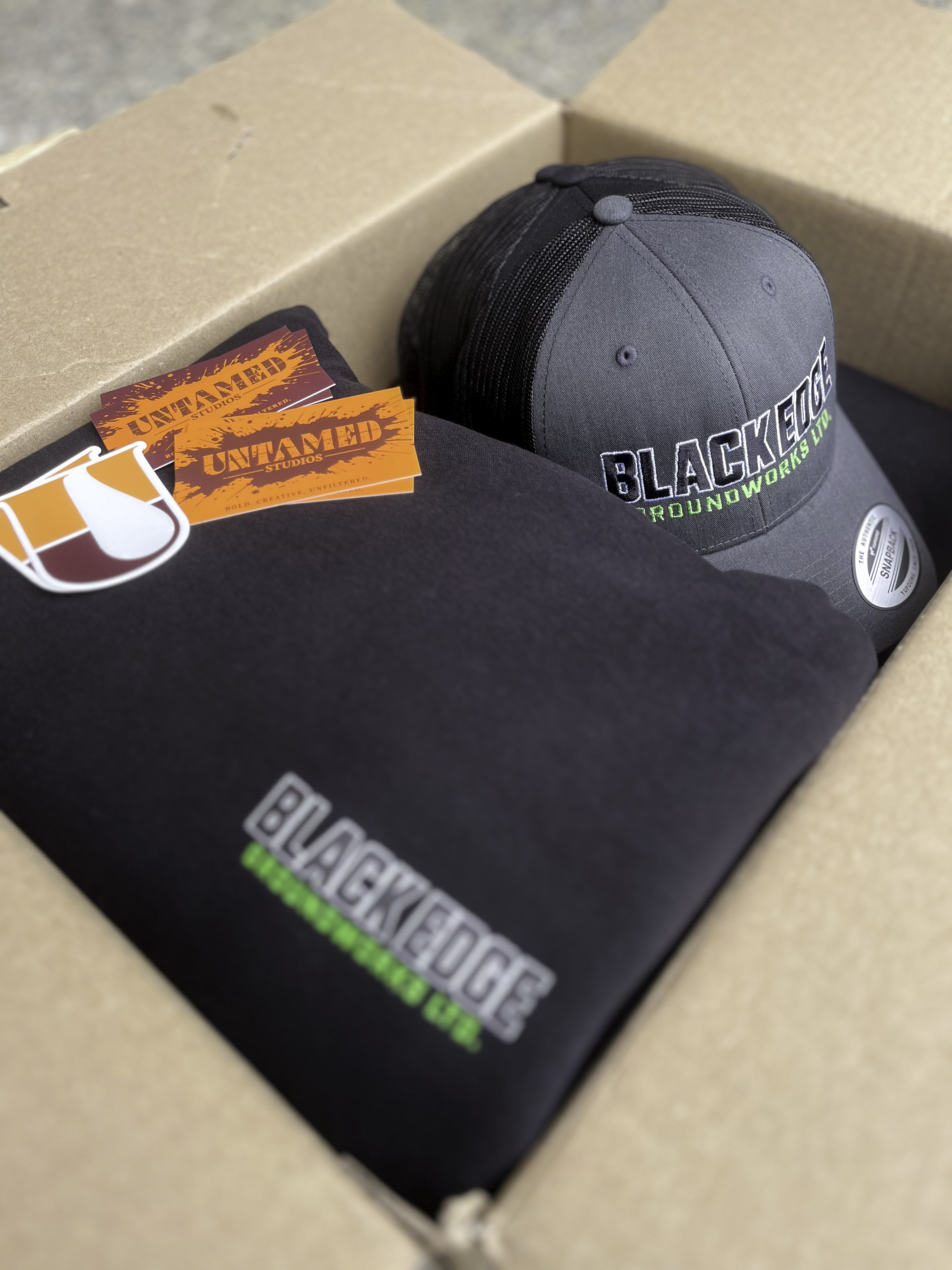 Black Edge Groundworks branded merch packed in a shipping box with Untamed Studios cards and stickers